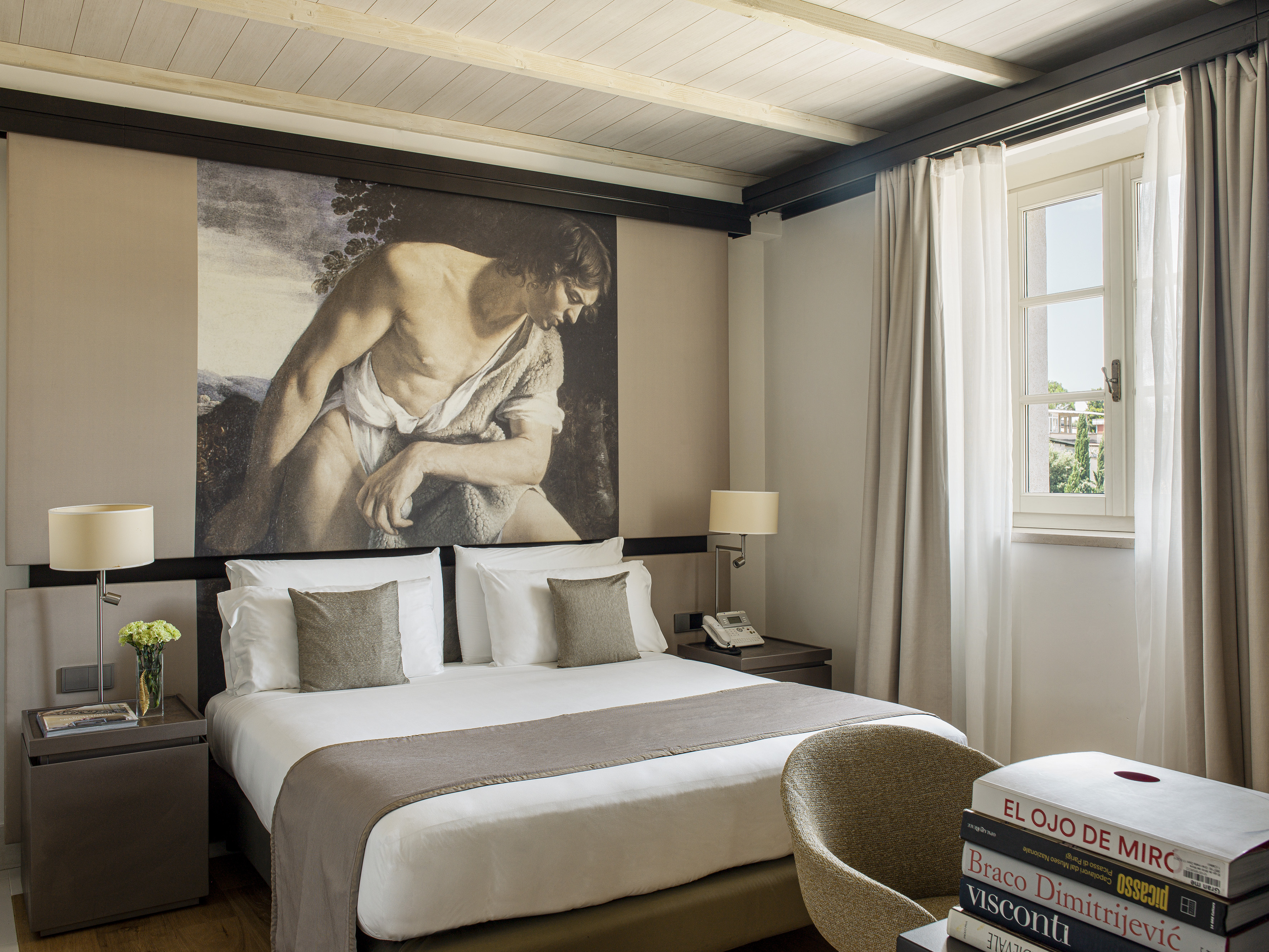 Elegant hotel bedroom with a large bed, classical art mural, and bright window.