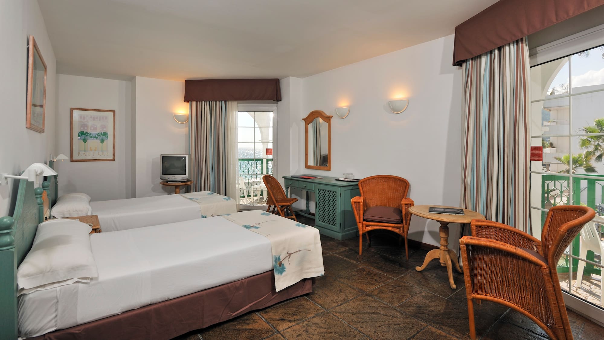 Coastal hotel room, twin beds, tiled floor, wicker furniture, balcony with sea view.