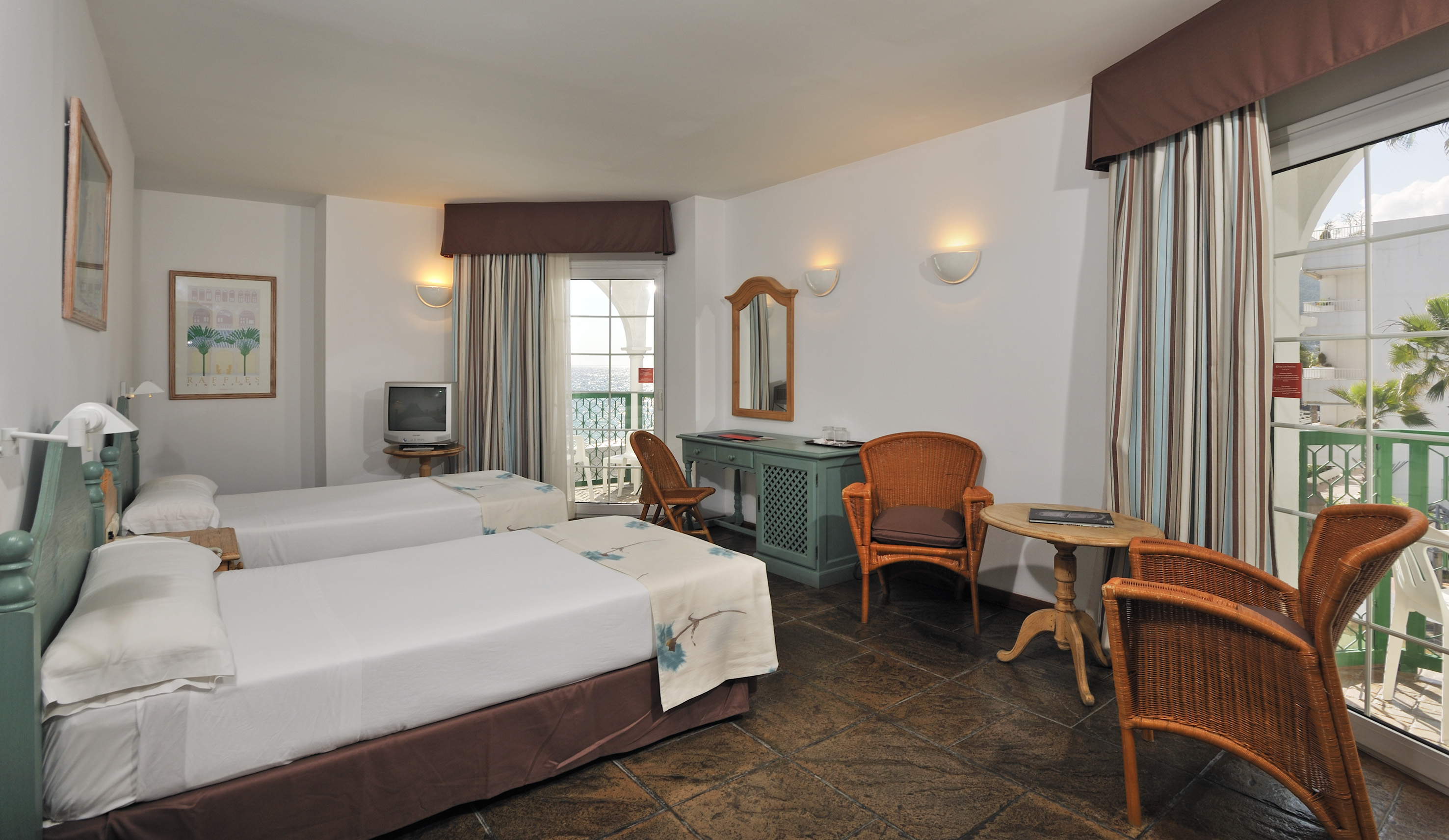 Coastal hotel room, twin beds, tiled floor, wicker furniture, balcony with sea view.