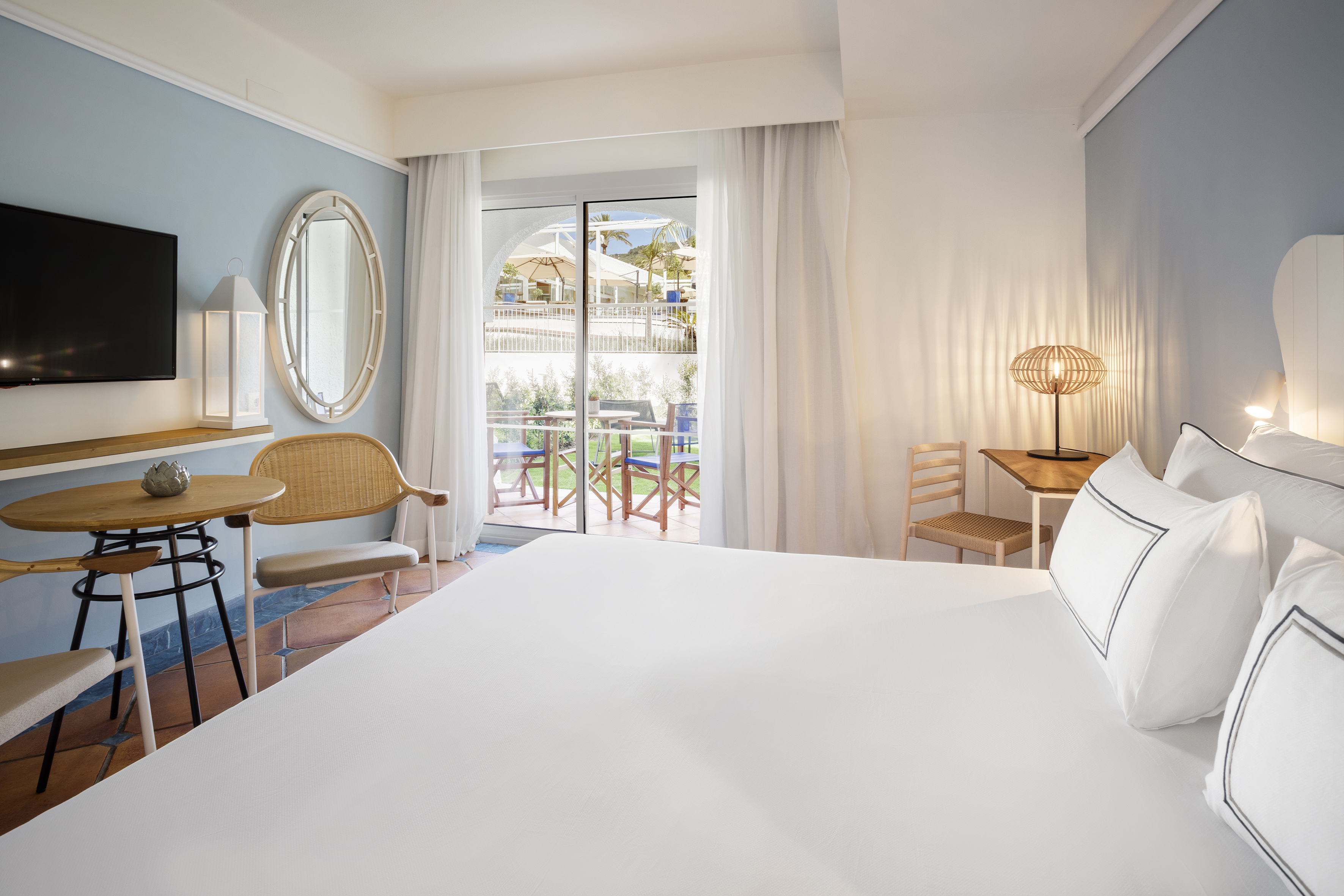 Hotel room with white bed, blue wall, and balcony with outdoor seating.