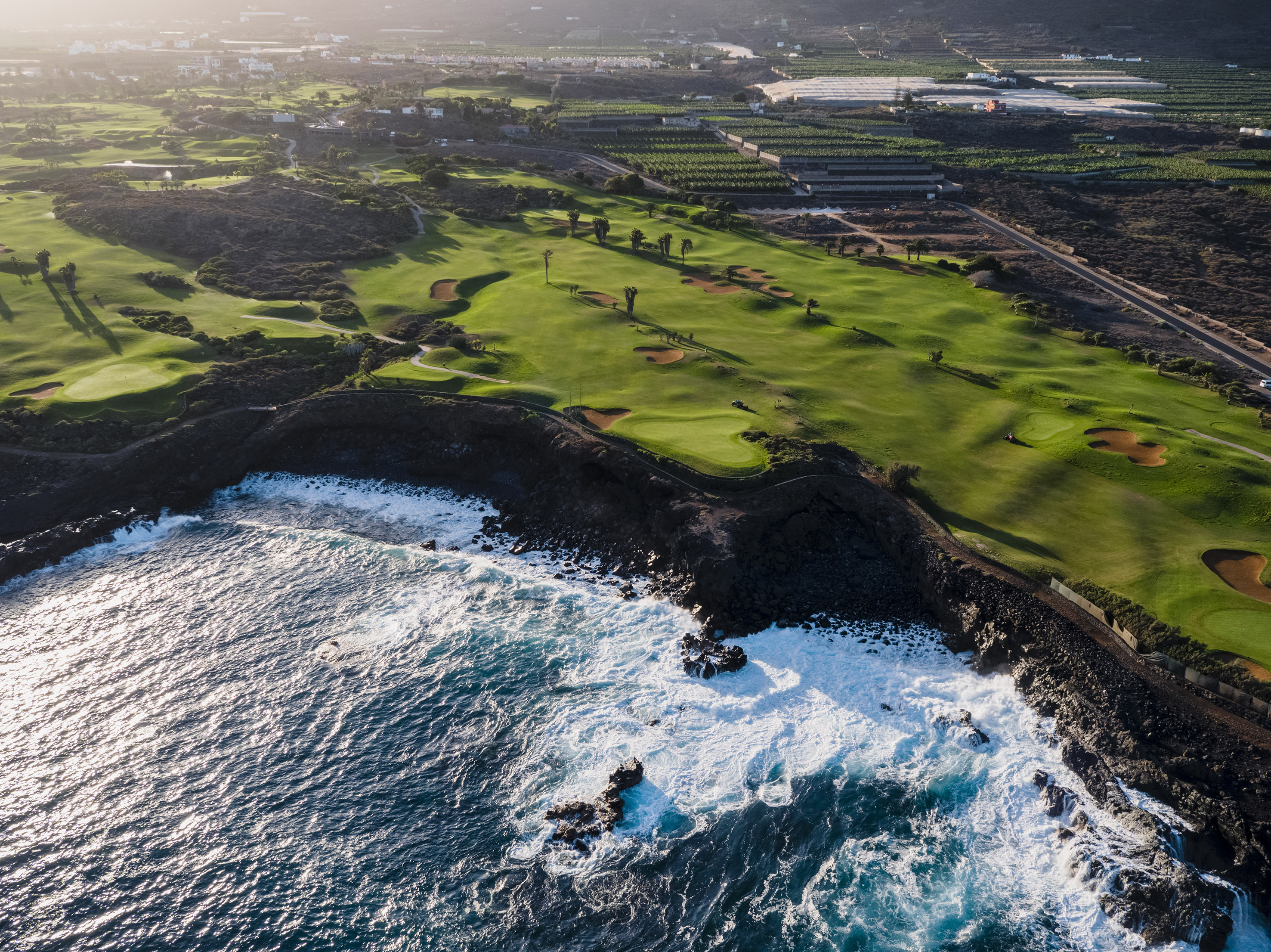 Aerial: Green golf course meets rocky coast with ocean waves and sand bunkers.