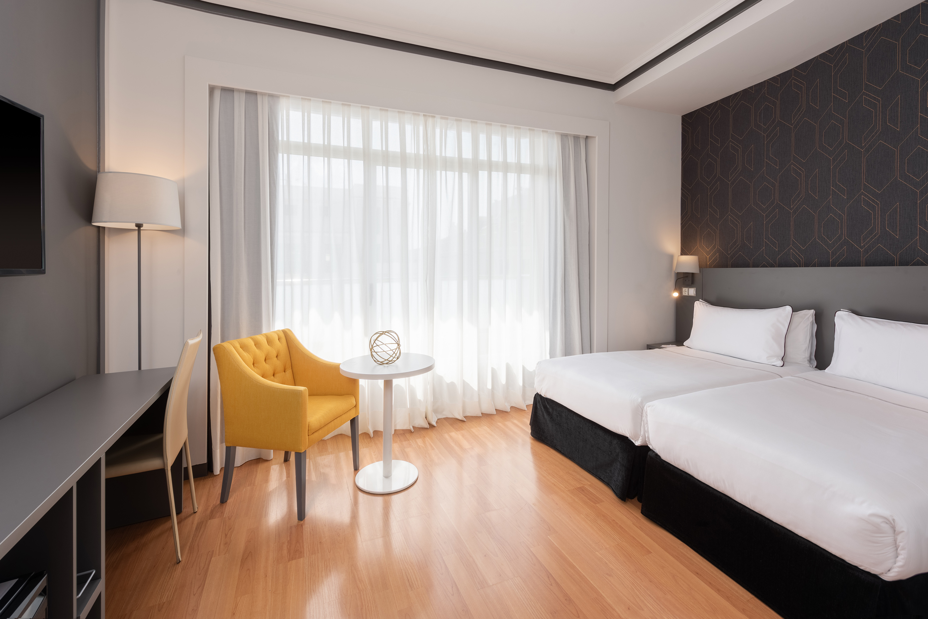 Modern hotel room with two twin beds, yellow armchair, desk, and sheer curtains.