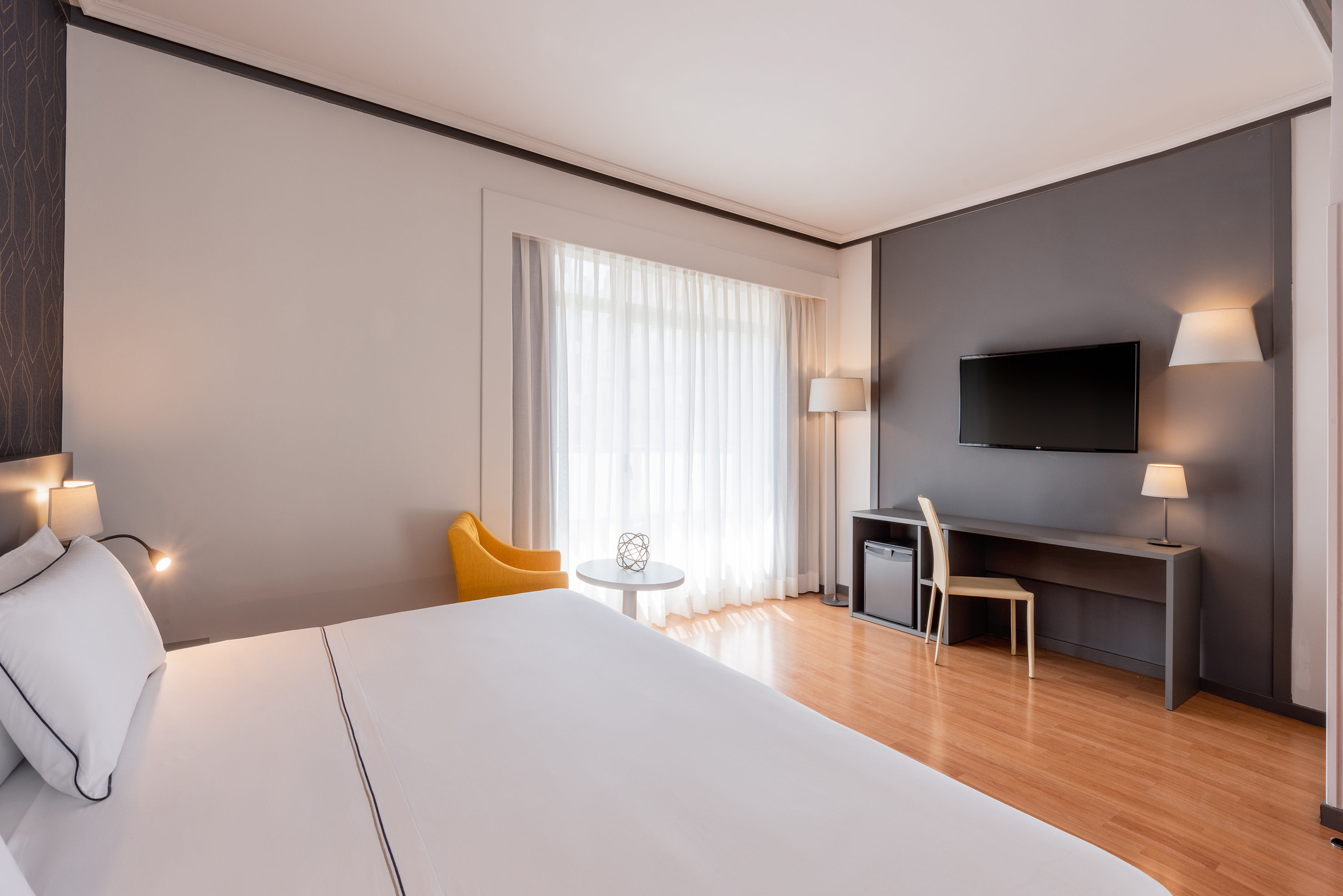 Modern hotel room with king bed, desk, TV, yellow armchair, wood floor.