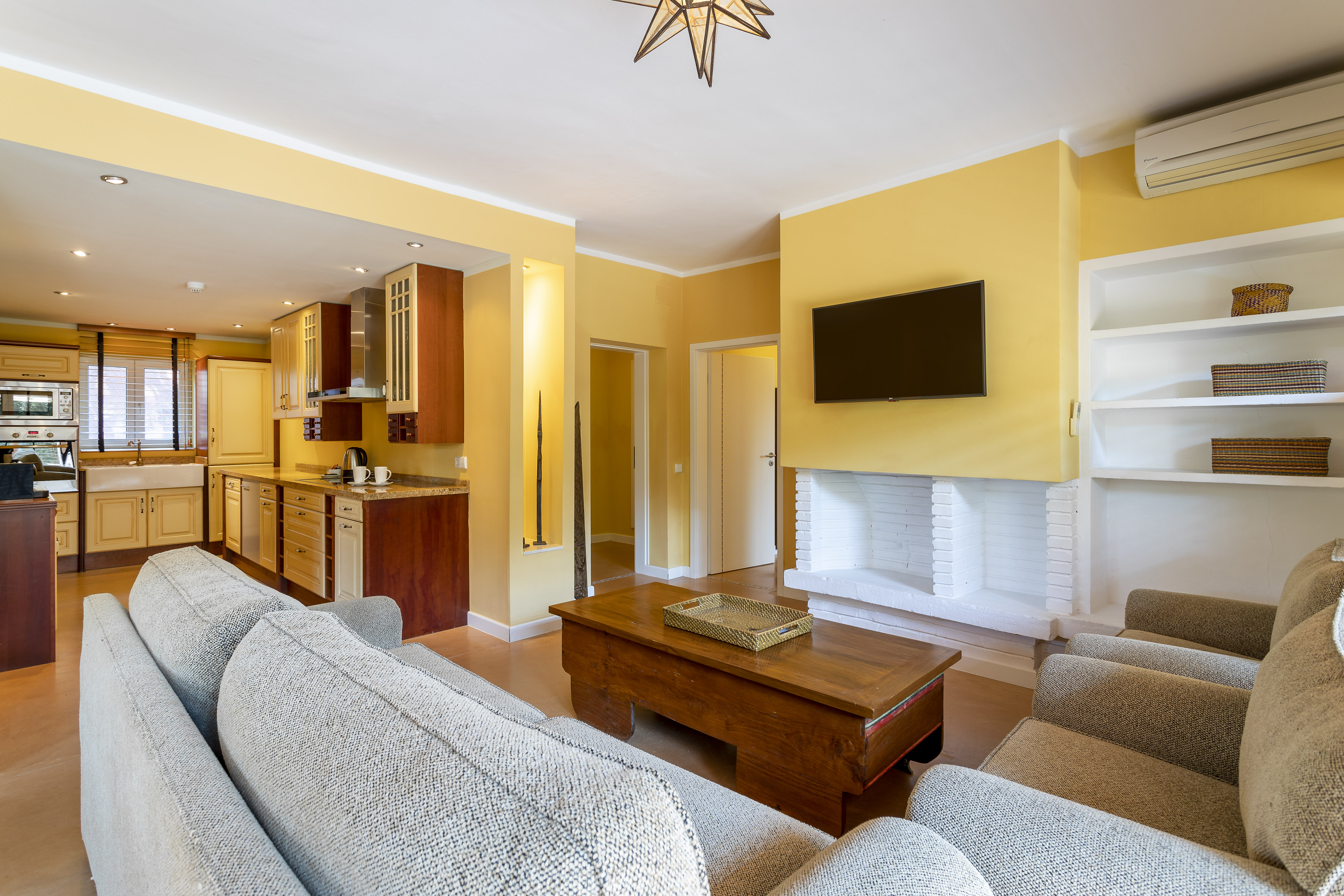 Yellow open-plan living room and kitchen with sofas, TV, and wooden cabinets.