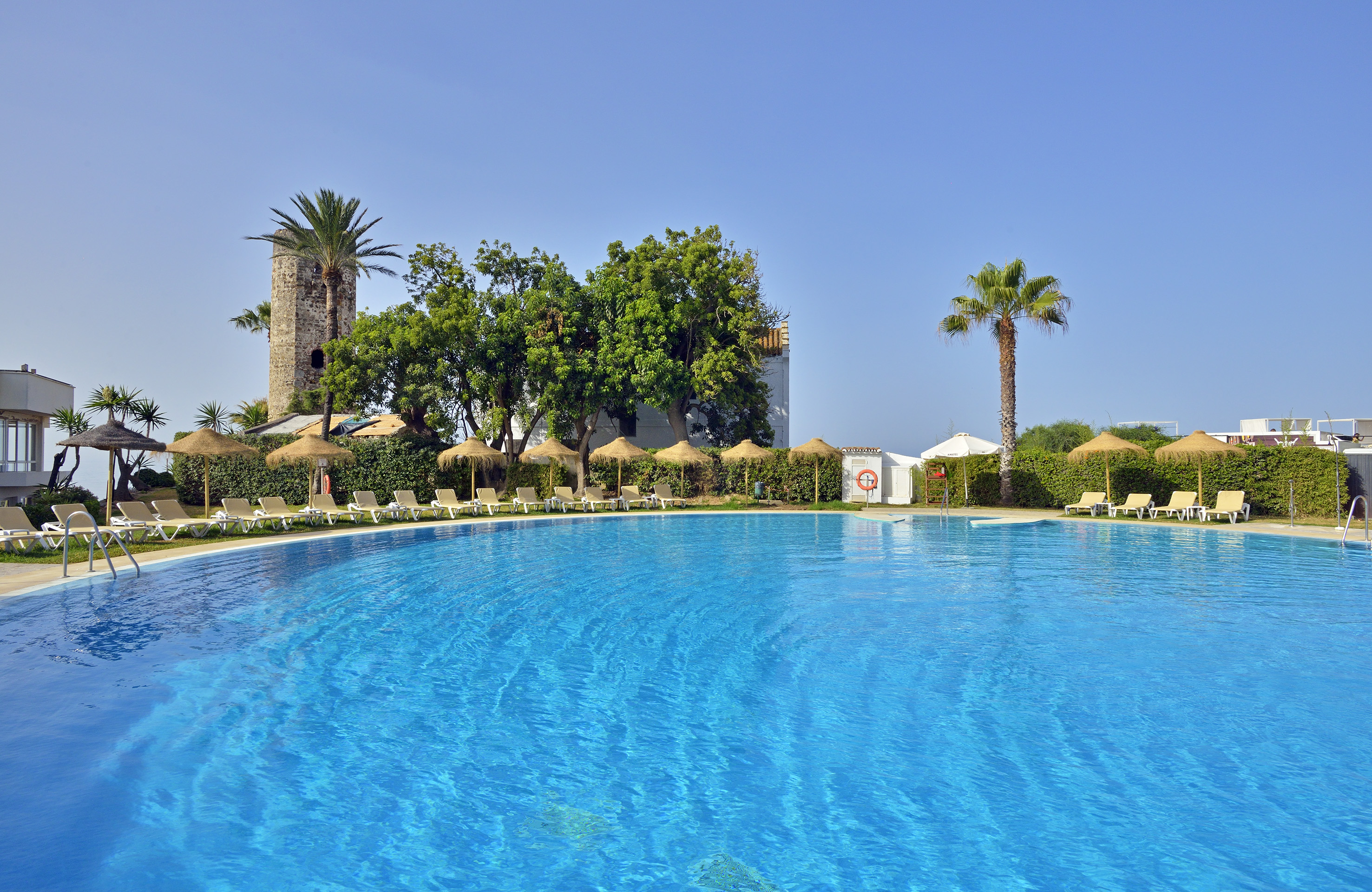 Resort swimming pool with sun loungers, palm trees, and a historic stone tower.