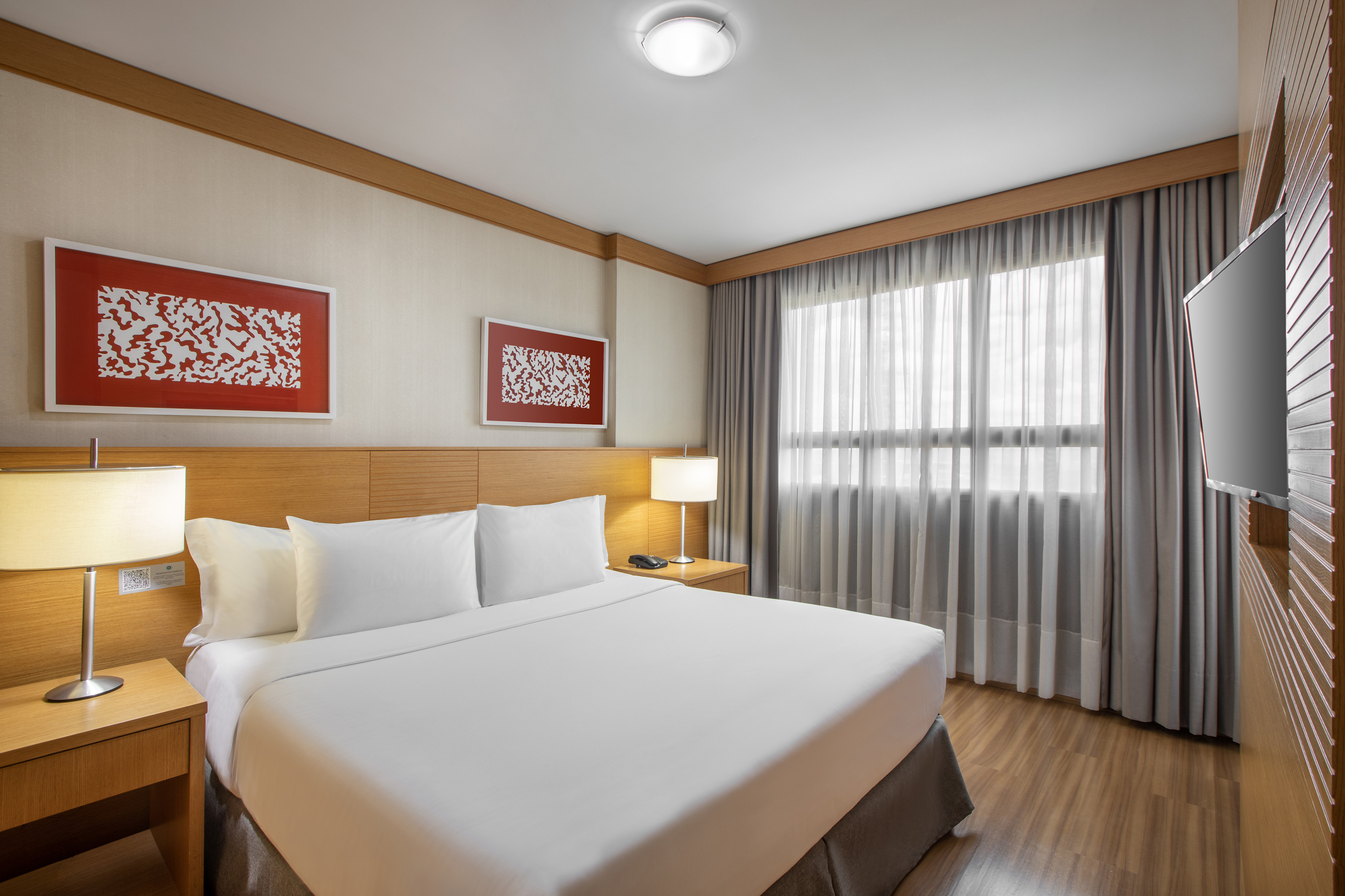 Modern hotel room with white bed, wooden headboard, window, and abstract art.