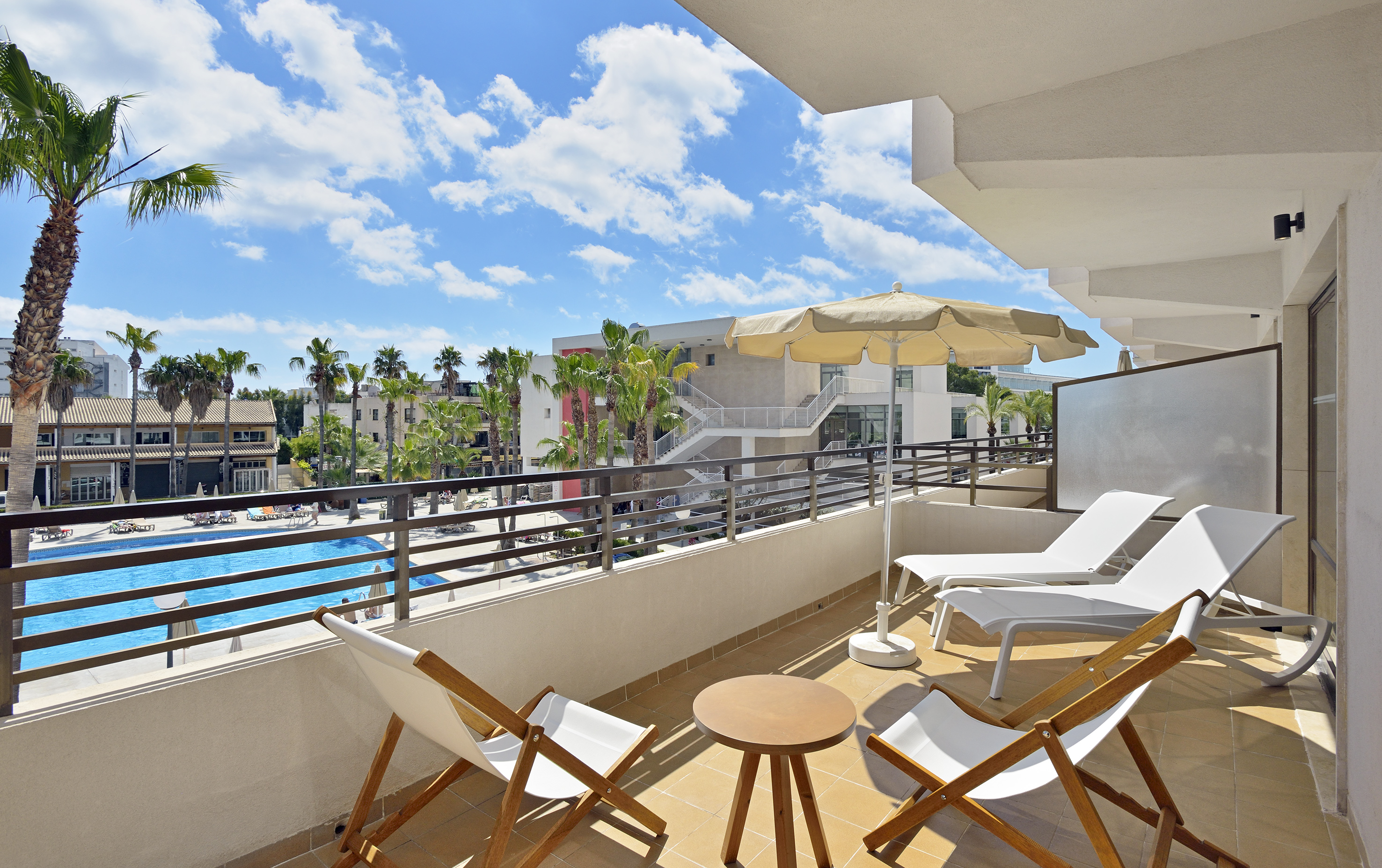 Hotel balcony with pool view, palm trees, chairs, and umbrella under a blue sky.