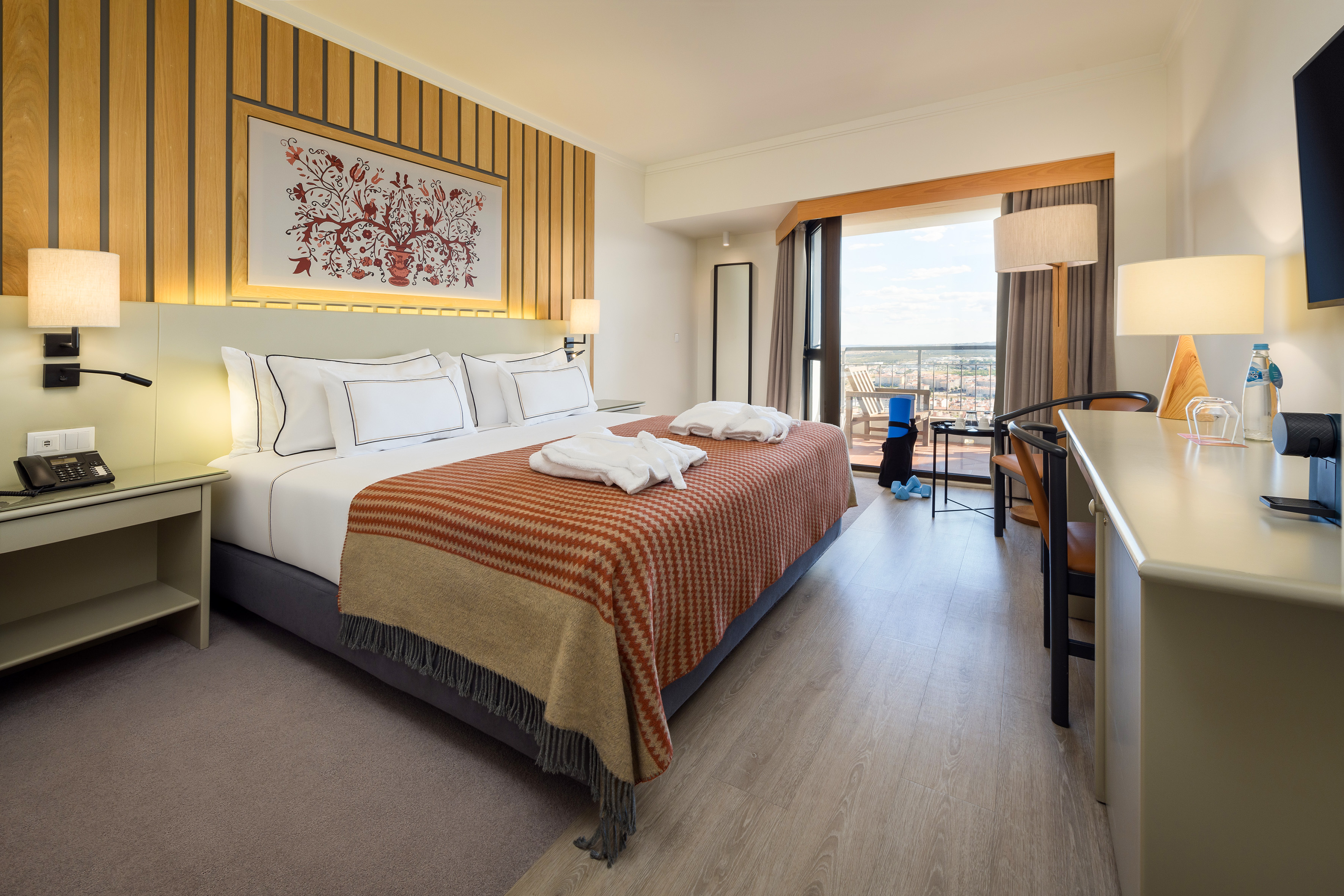 Modern hotel room with king bed, city view balcony, desk, and exercise gear.