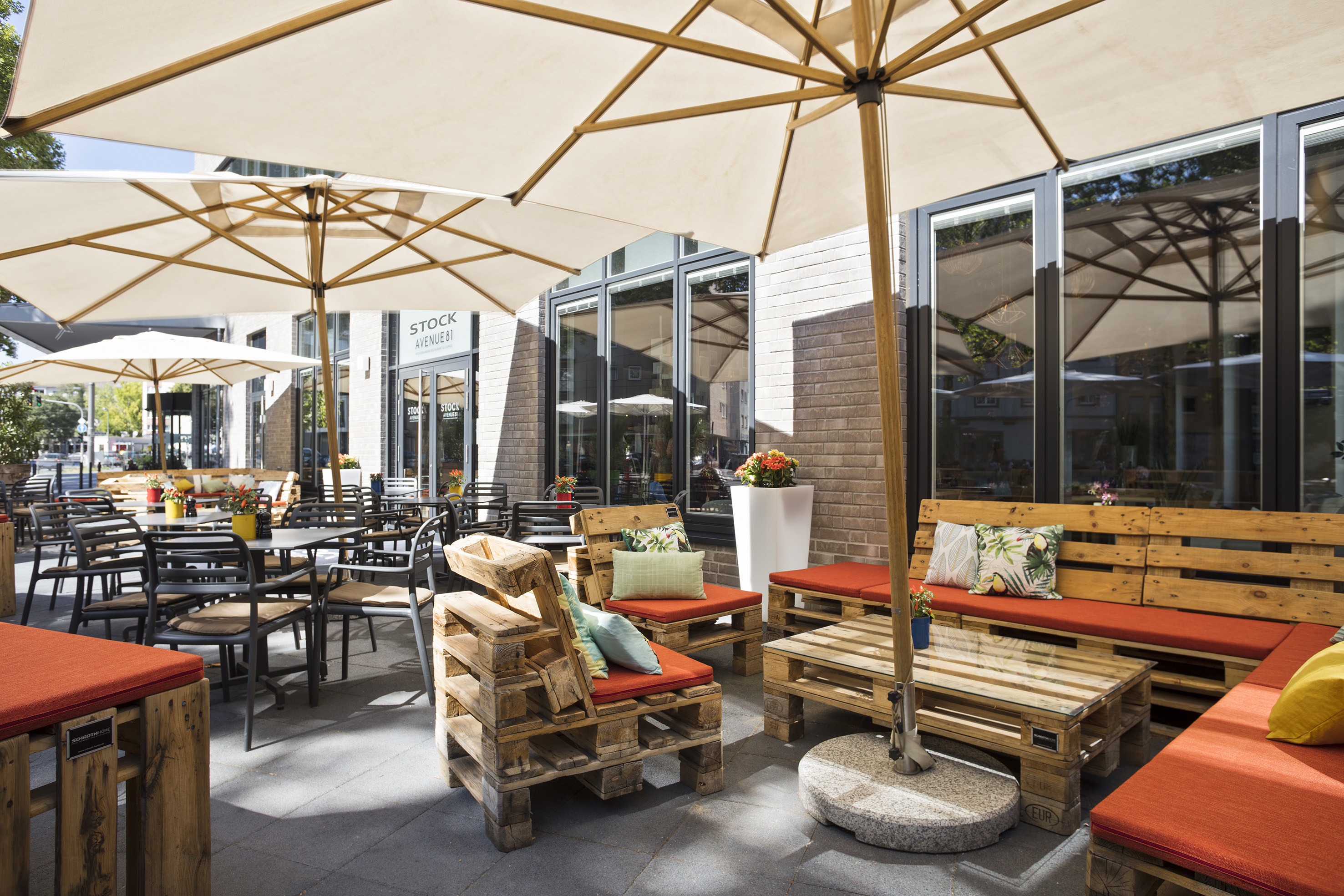Outdoor cafe terrace with pallet seating, orange cushions, and large umbrellas.
