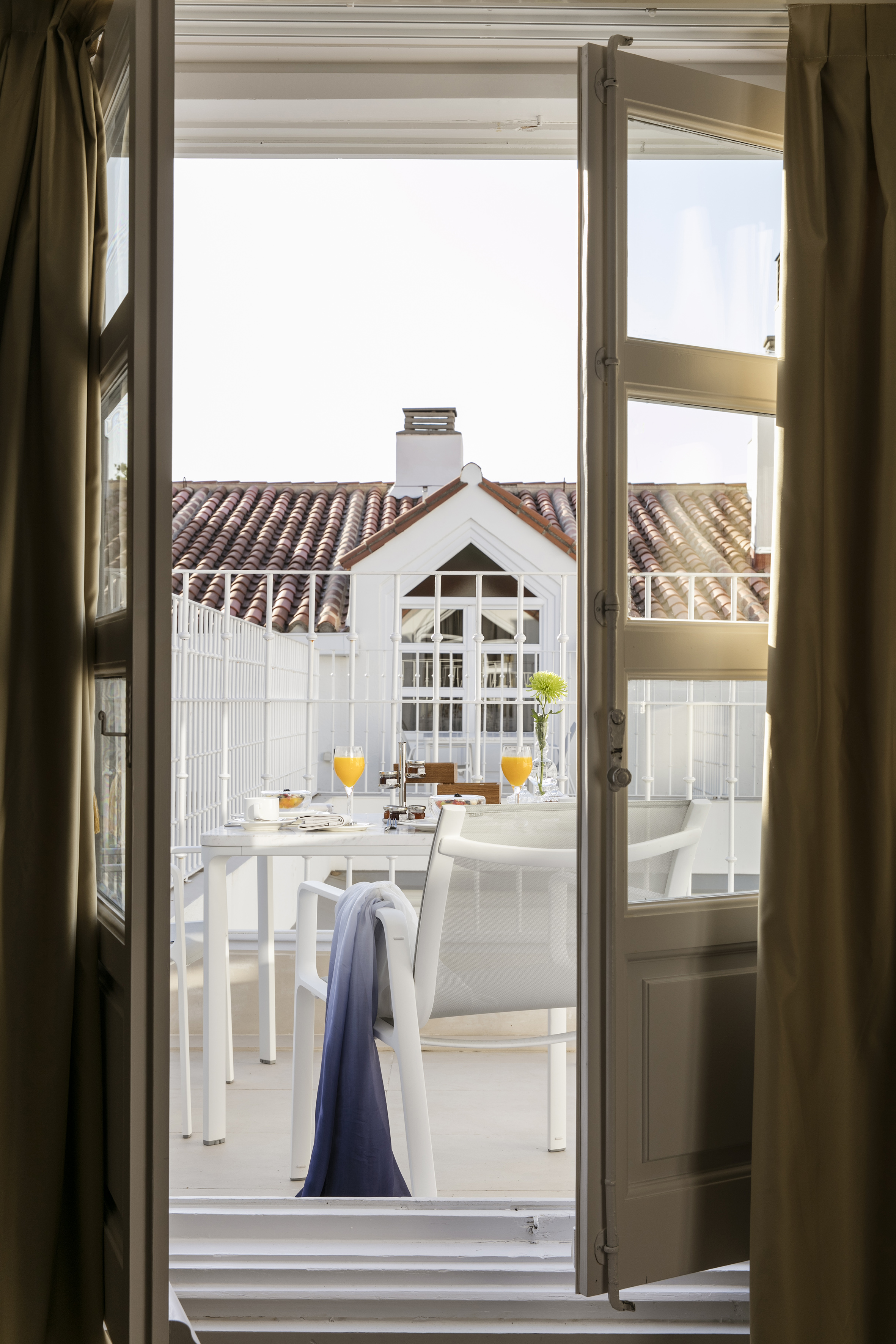 Open balcony doors to outdoor breakfast table with juice, white chairs, tiled roof view.