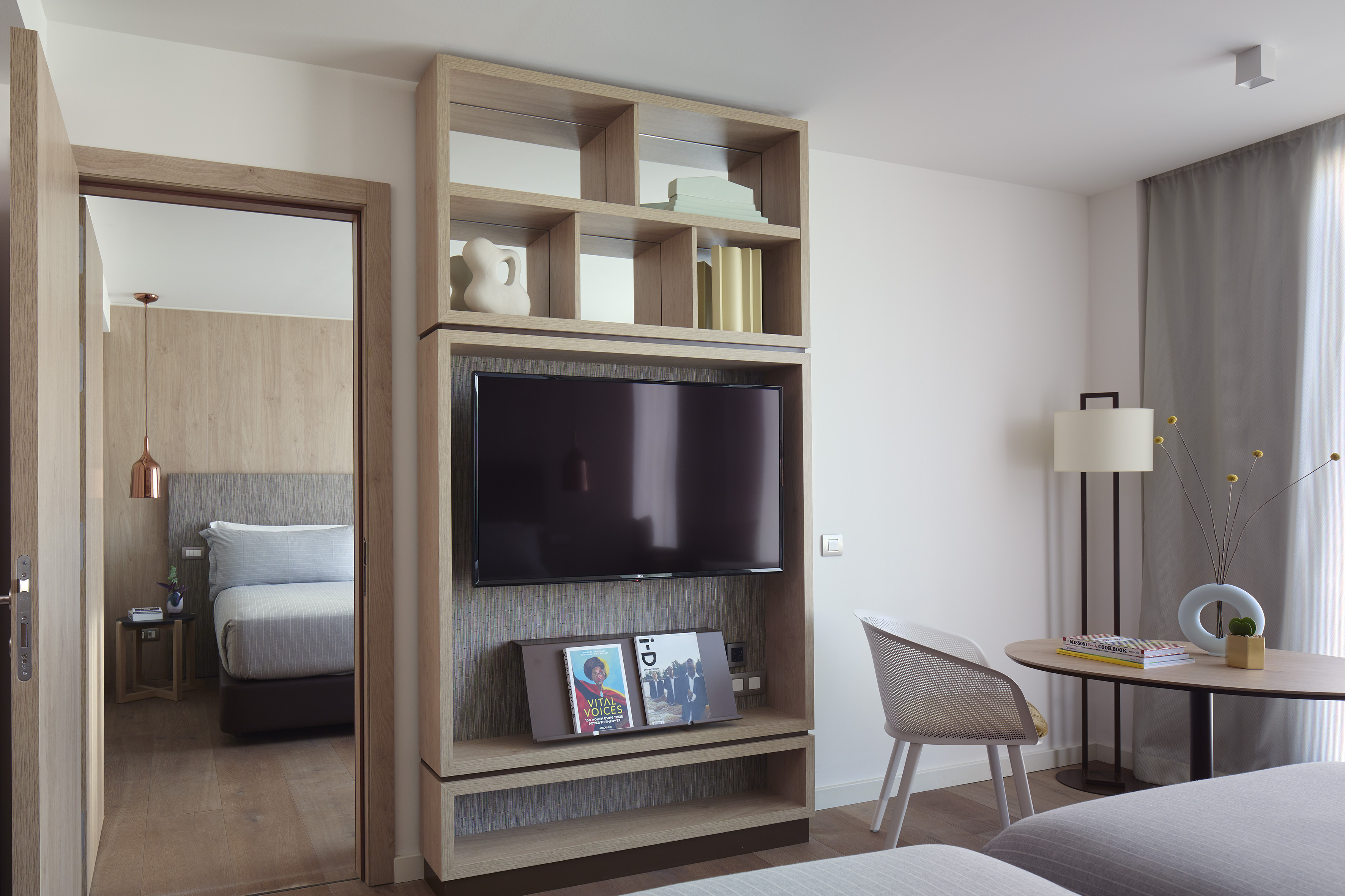 Modern hotel living area with wooden TV unit, decor shelves, and adjacent bedroom.