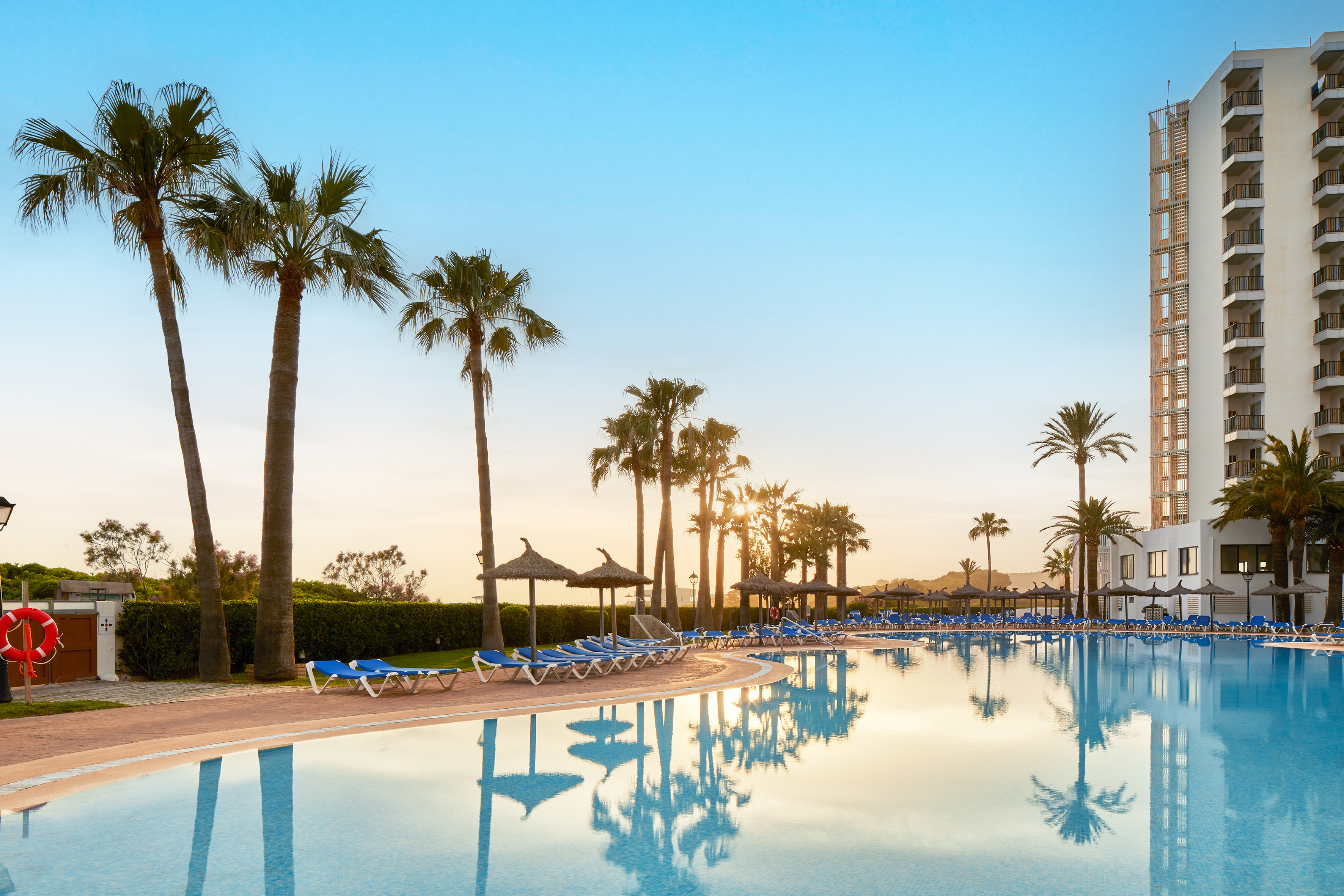 Piscina de hotel al atardecer con palmeras, tumbonas y edificio moderno.