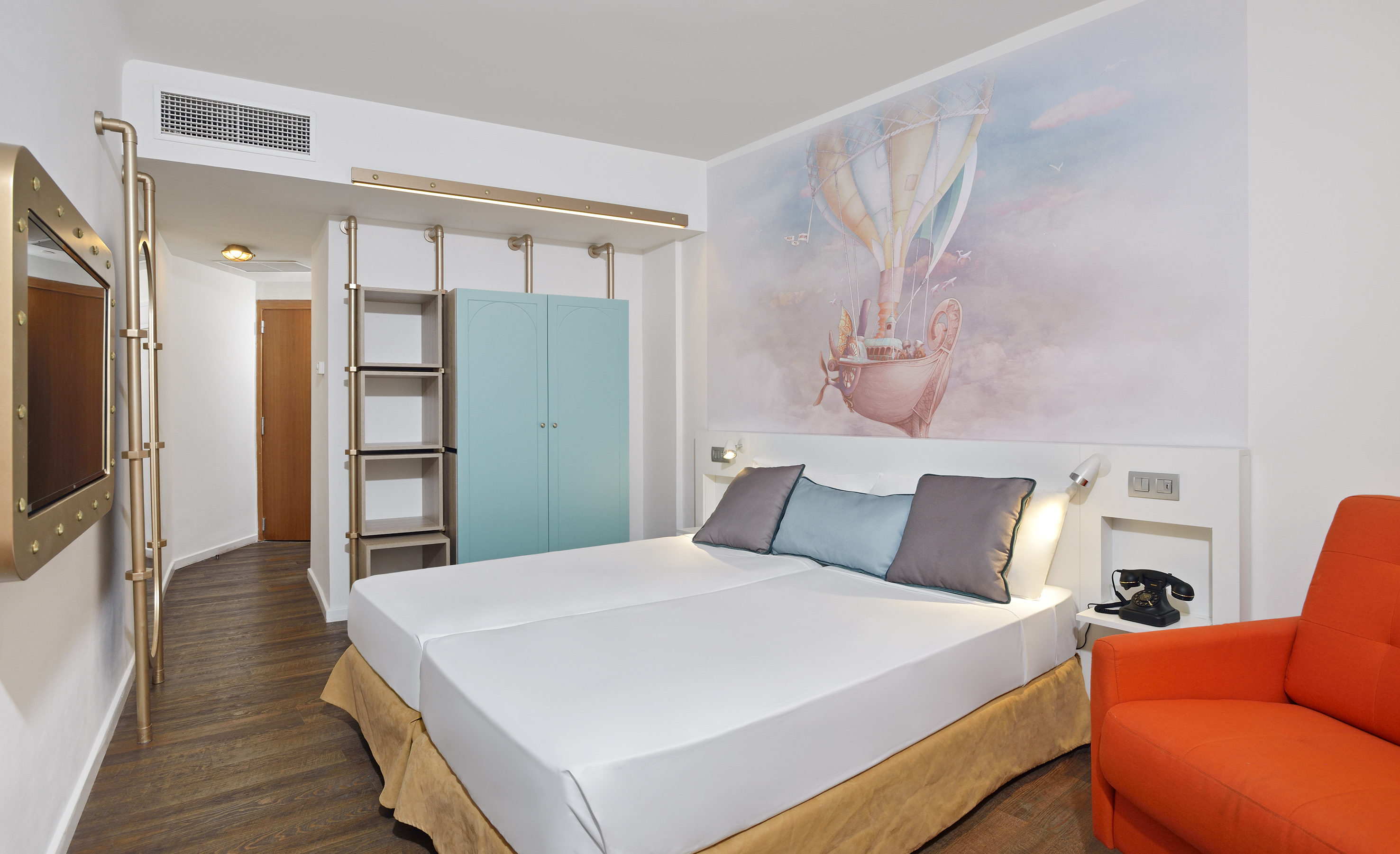 Modern hotel room: hot air balloon mural, twin beds, orange chair, blue wardrobe.