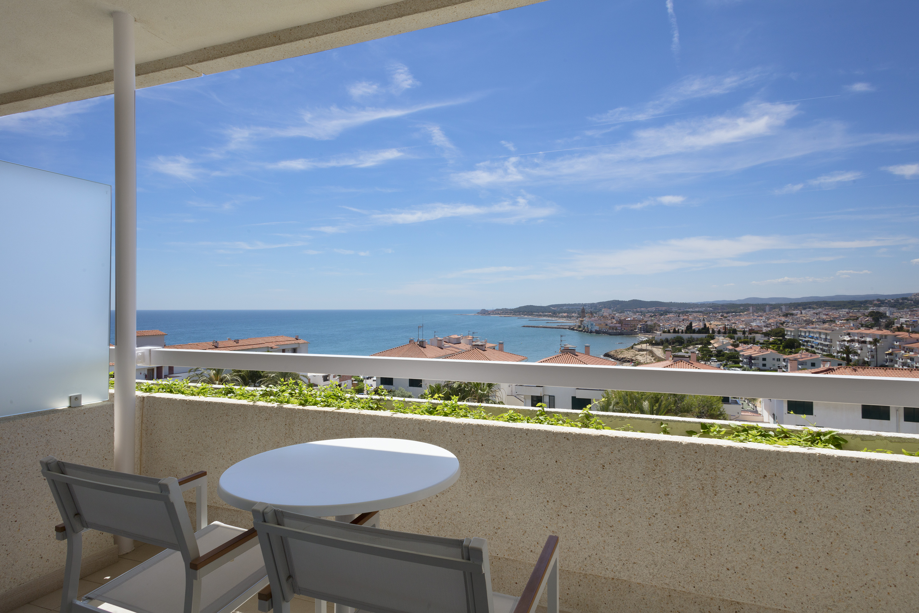Balcony with outdoor seating, overlooking blue sea and coastal city.