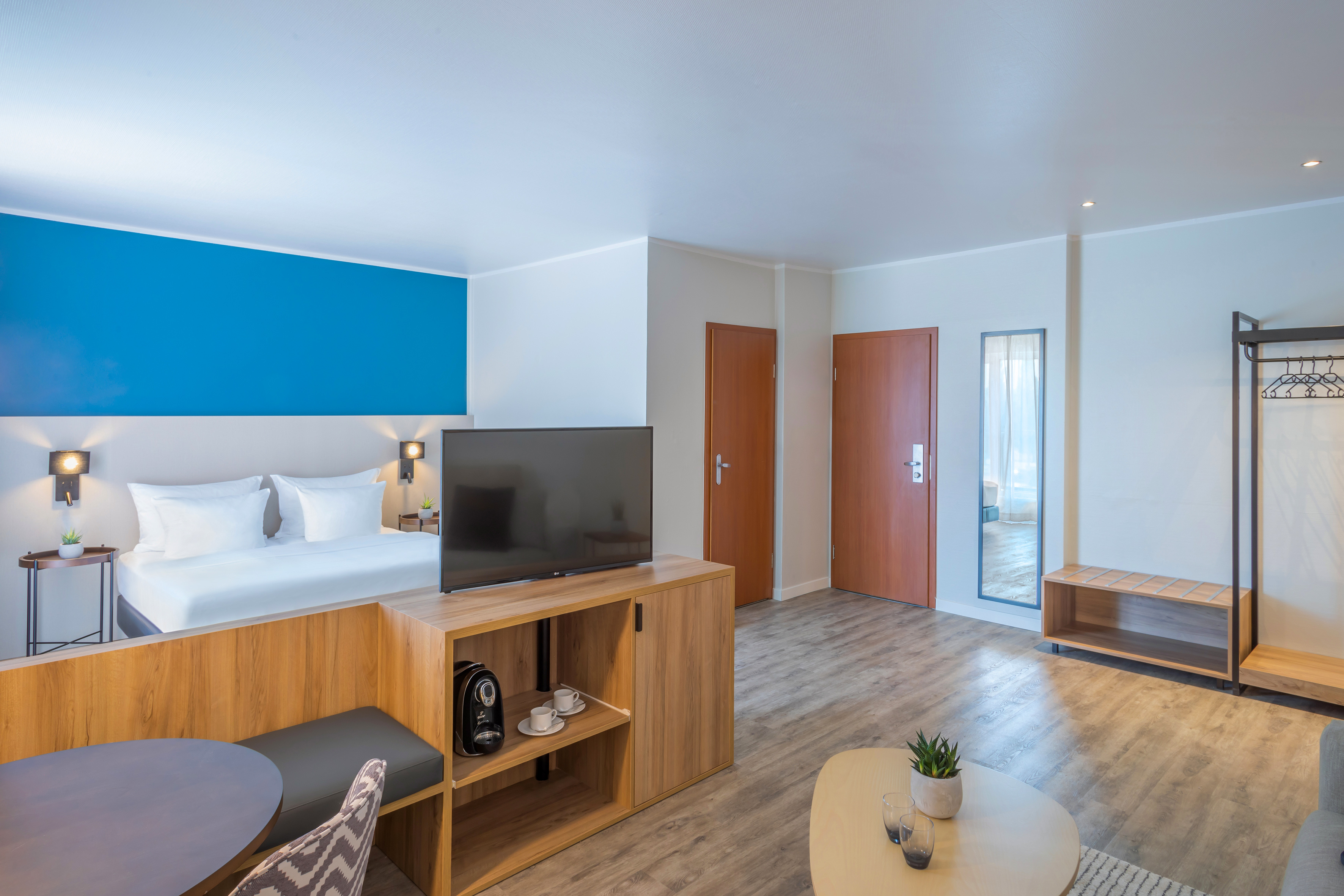 Modern hotel room suite with bed, TV, blue accent wall, coffee machine, and dining.