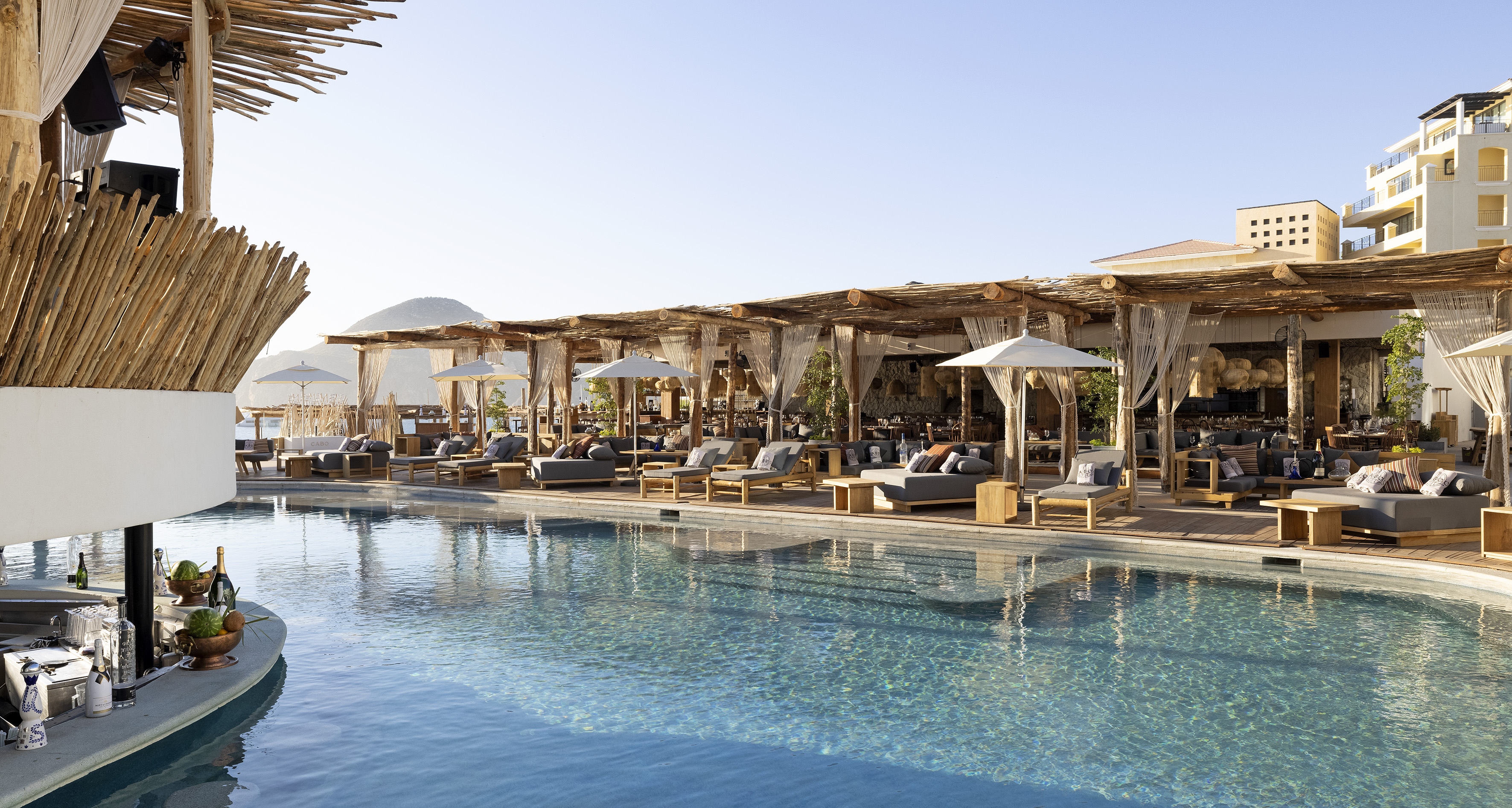 Cabo luxury resort pool lounge with daybeds, wooden pergola, and ocean.