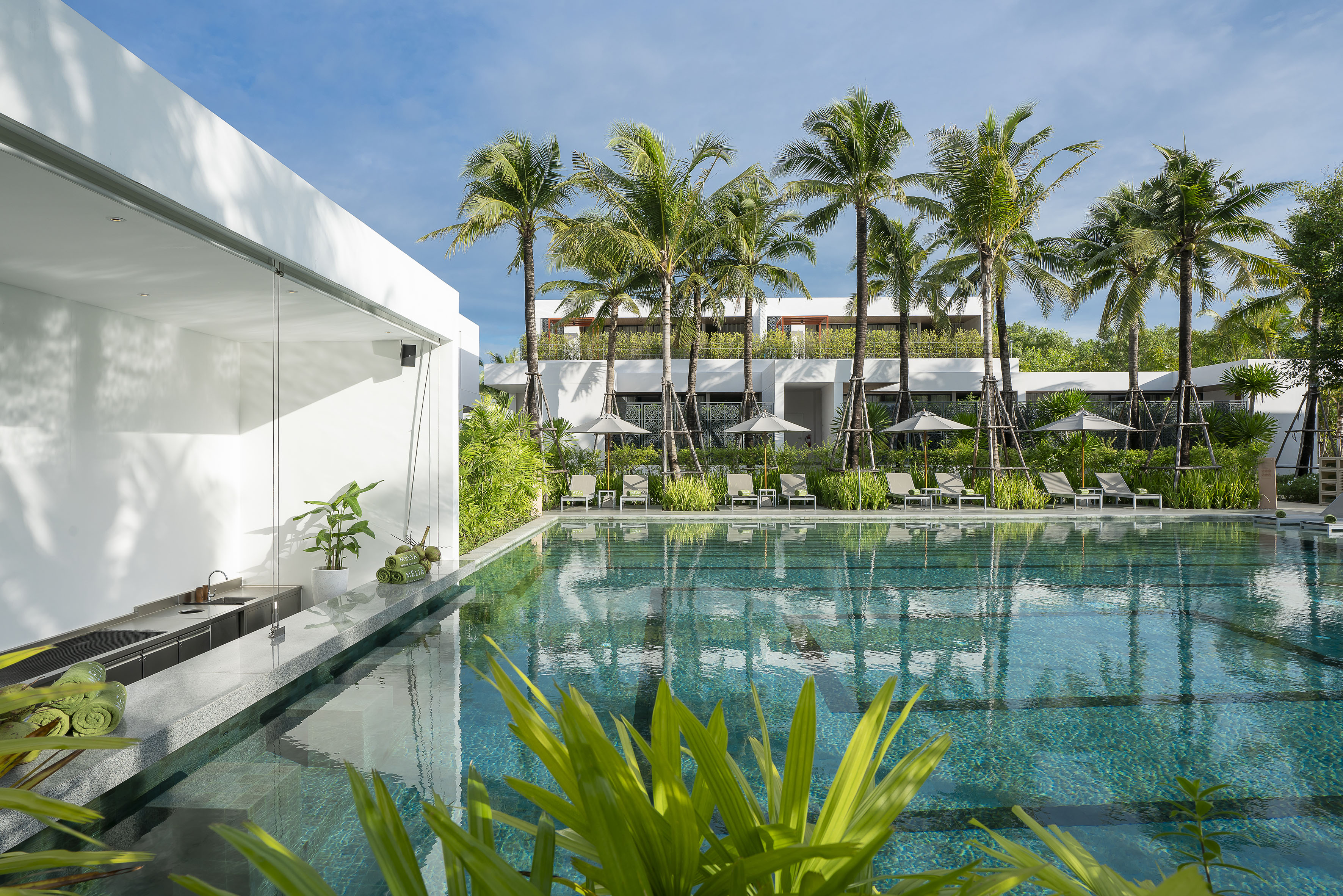 Tropical resort swimming pool with palm trees, lounge chairs, and poolside bar.