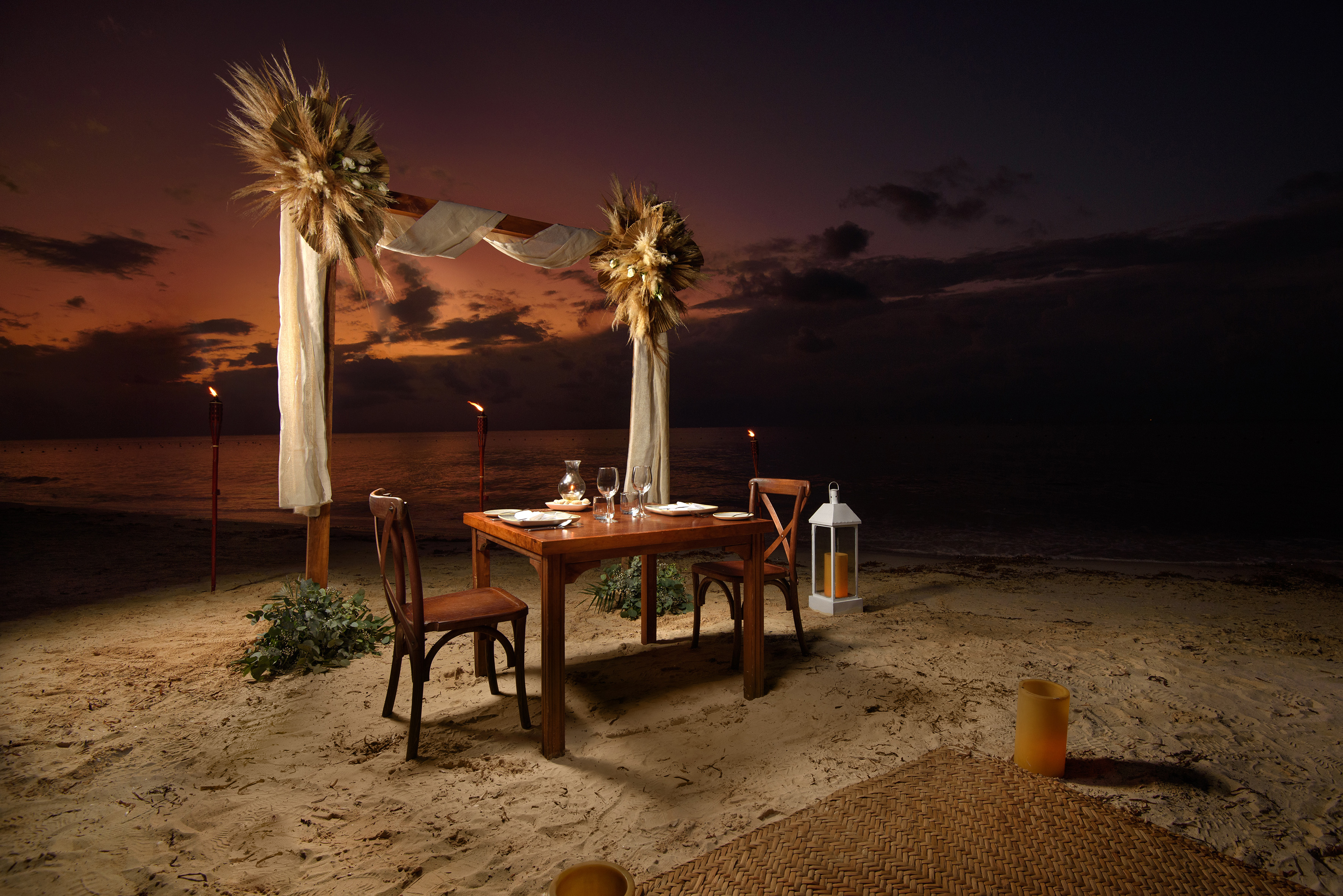 Romantic beach dinner setup for two at sunset with arch and tiki torches.