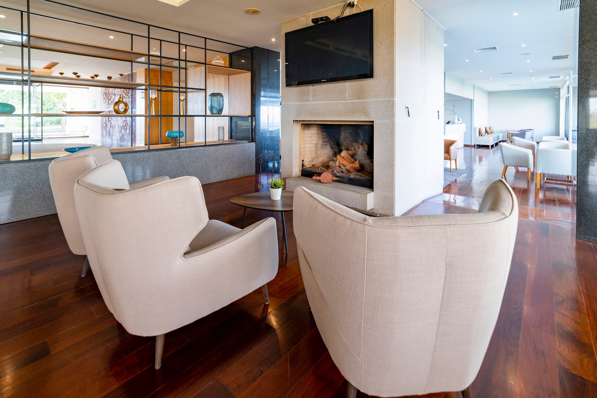 Hotel lounge with fireplace, TV, cream armchairs, and modern wood floor.