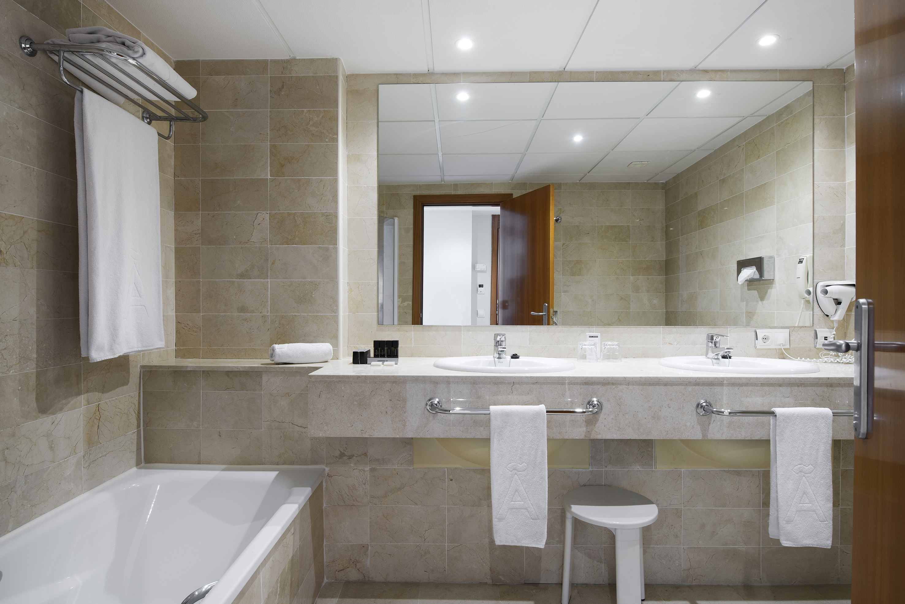 Hotel bathroom with dual sinks, bathtub, large mirror, and light beige tiles.