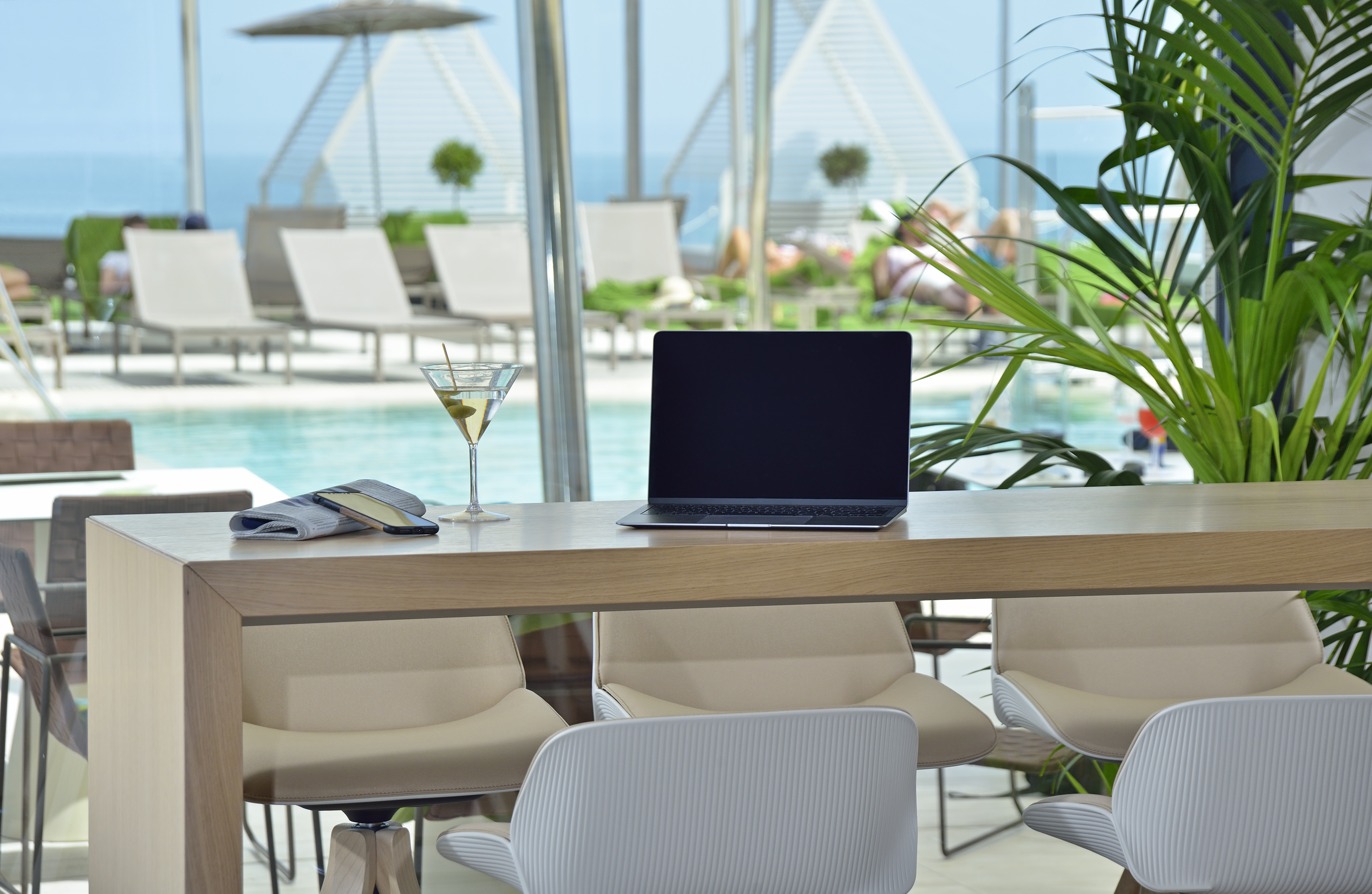 Modern bar with laptop, martini, overlooking a luxurious resort pool.