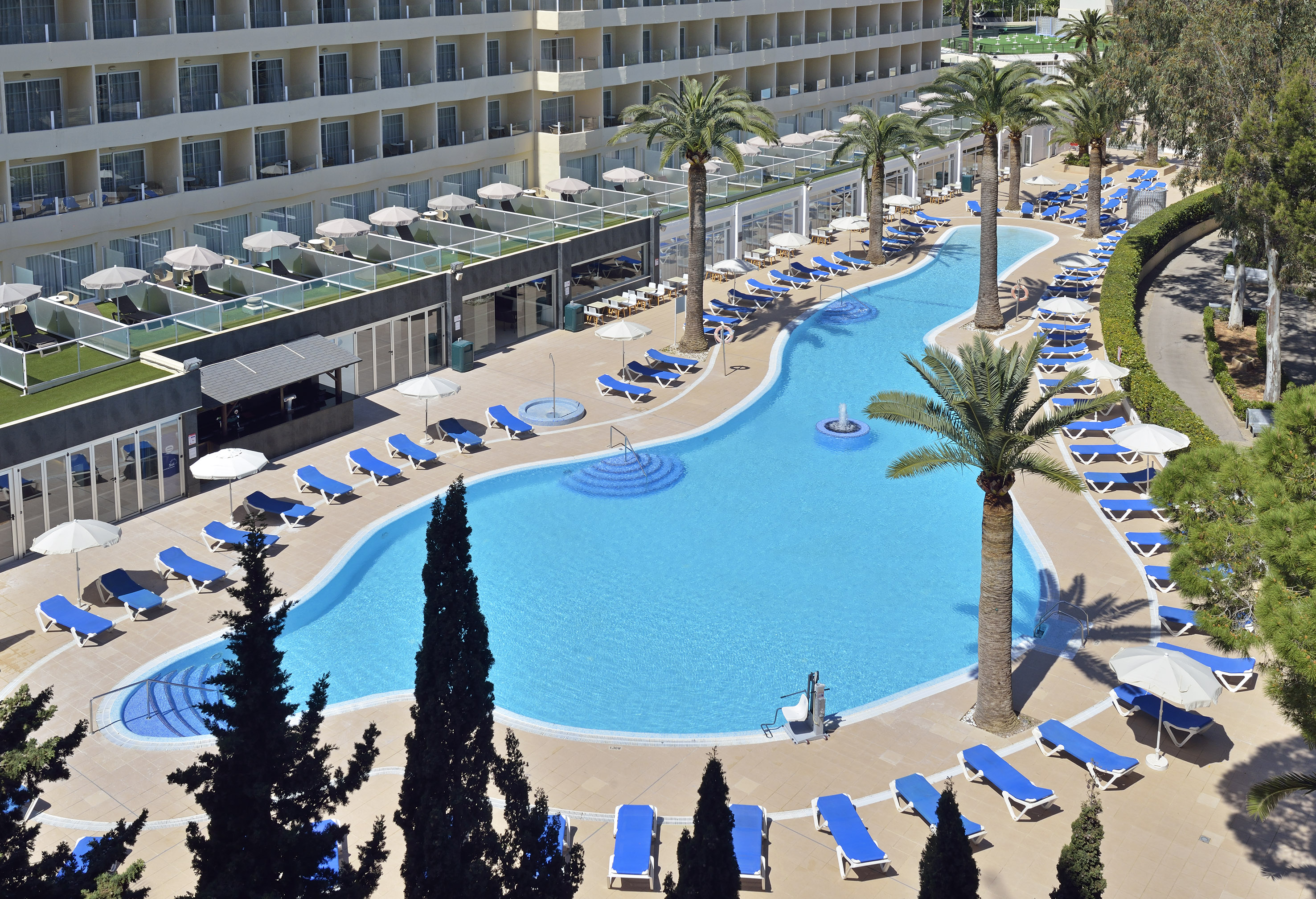 Large hotel pool with blue lounge chairs, umbrellas, and tropical palm trees.