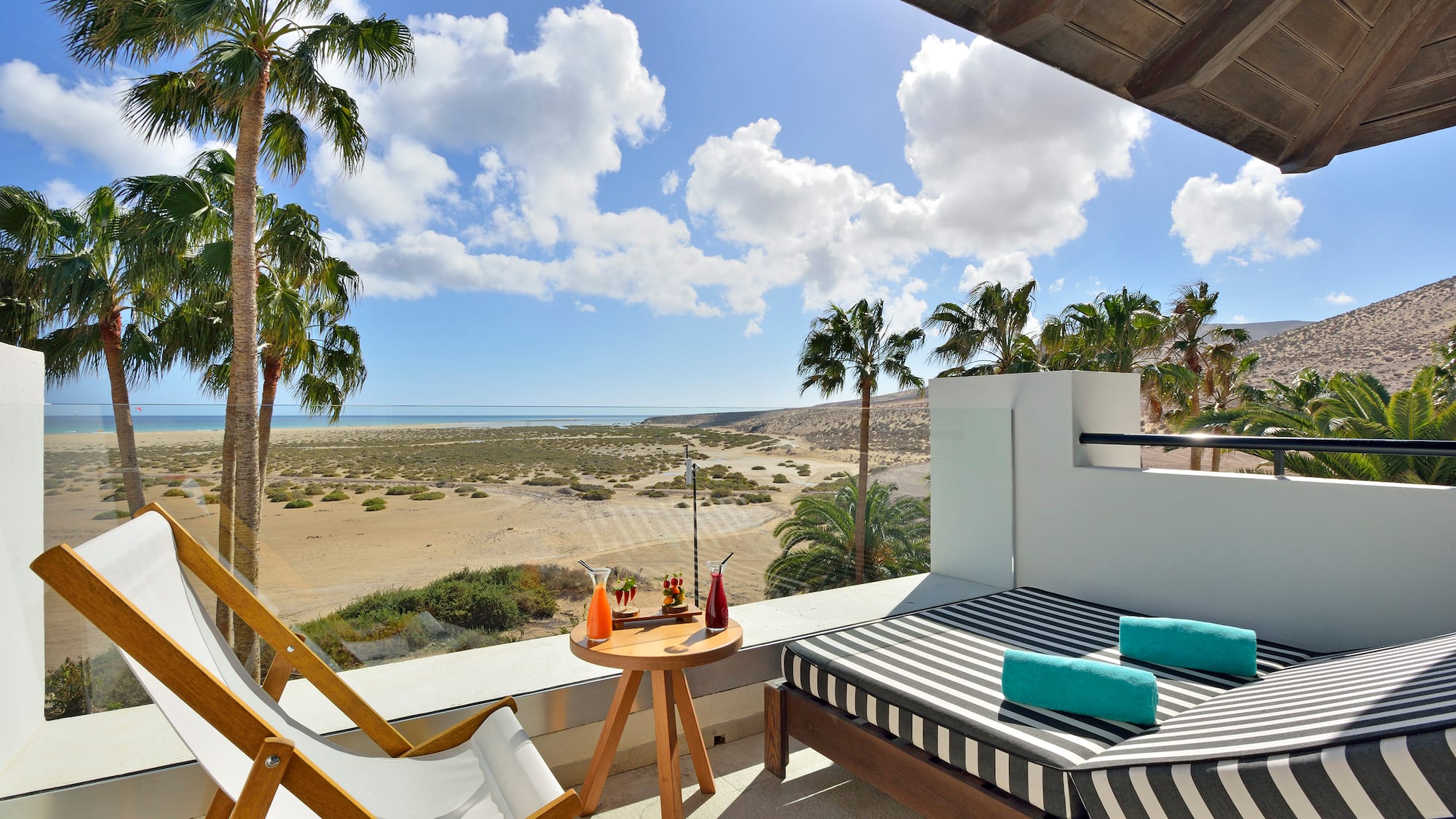 a deck with a bed and a table on a balcony overlooking a sandy beach