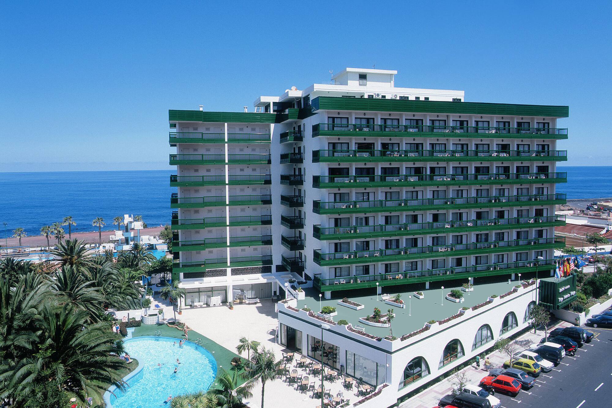 Green and white oceanfront hotel with pool, palm trees, and sea view.