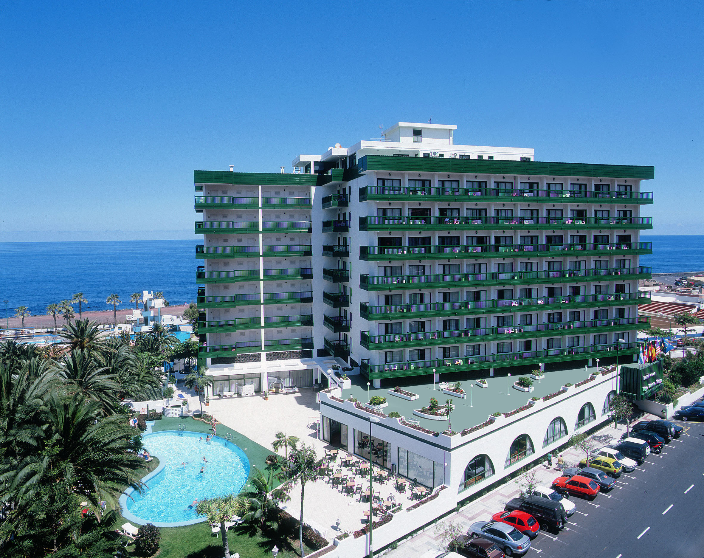 Green and white oceanfront hotel with pool, palm trees, and sea view.