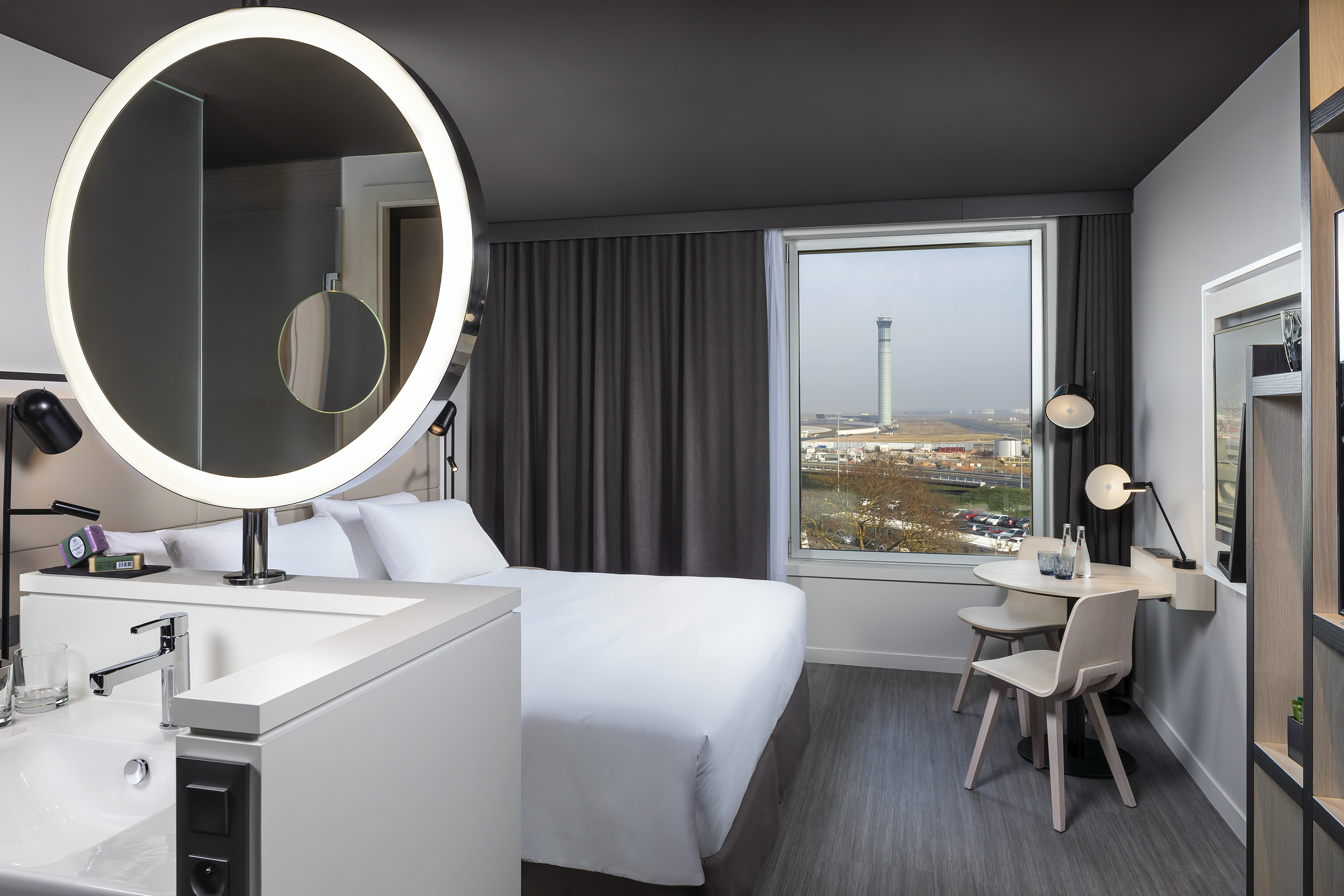 Airport hotel room with runway view, modern bed, backlit mirror, and desk.