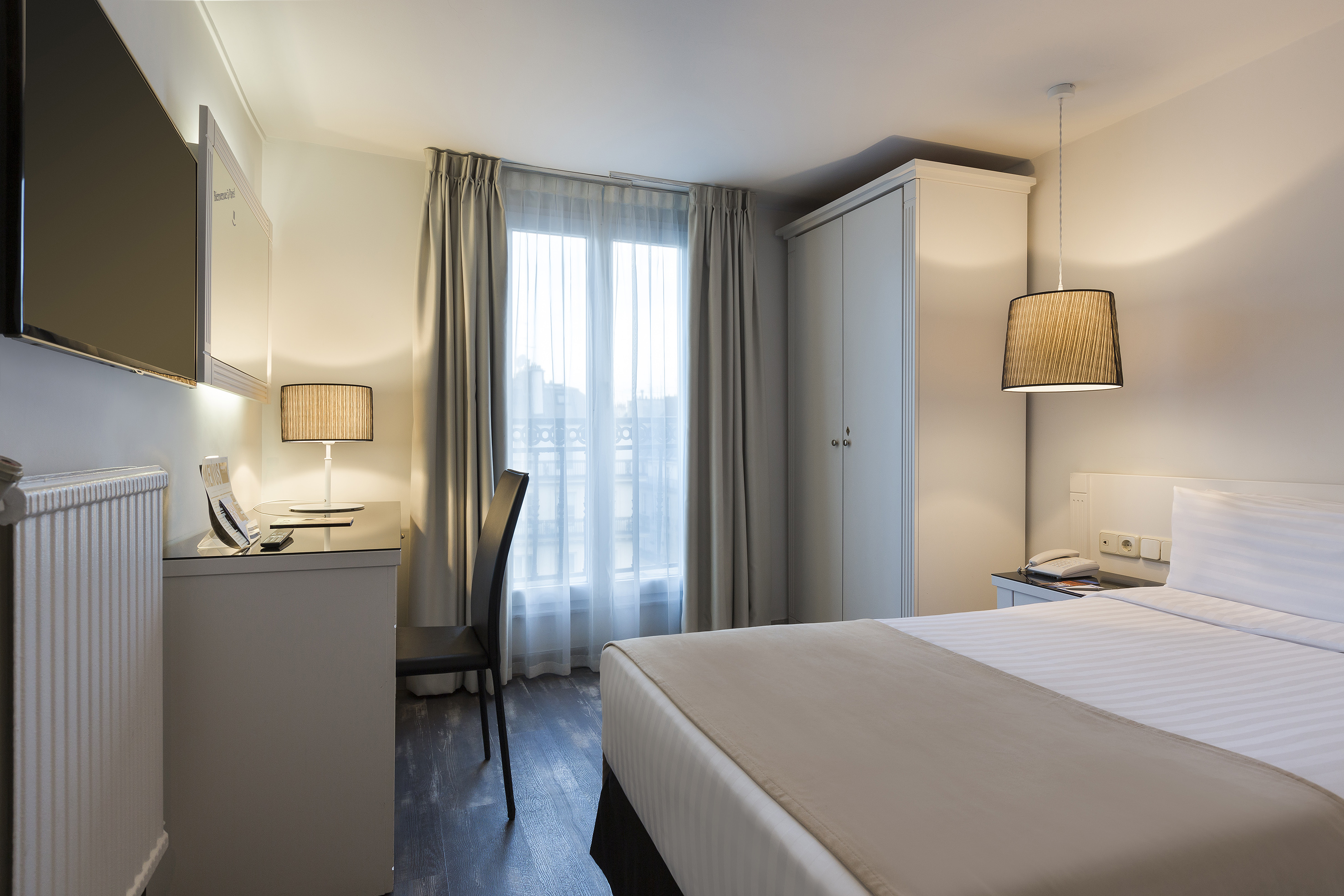 Bright hotel bedroom with bed, desk, TV, wardrobe, and window with curtains.