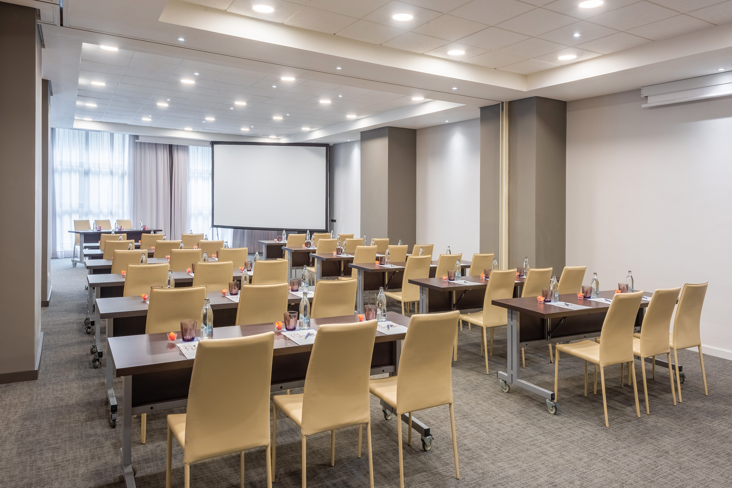 Modern conference room with projector screen, rows of tables, and beige chairs.