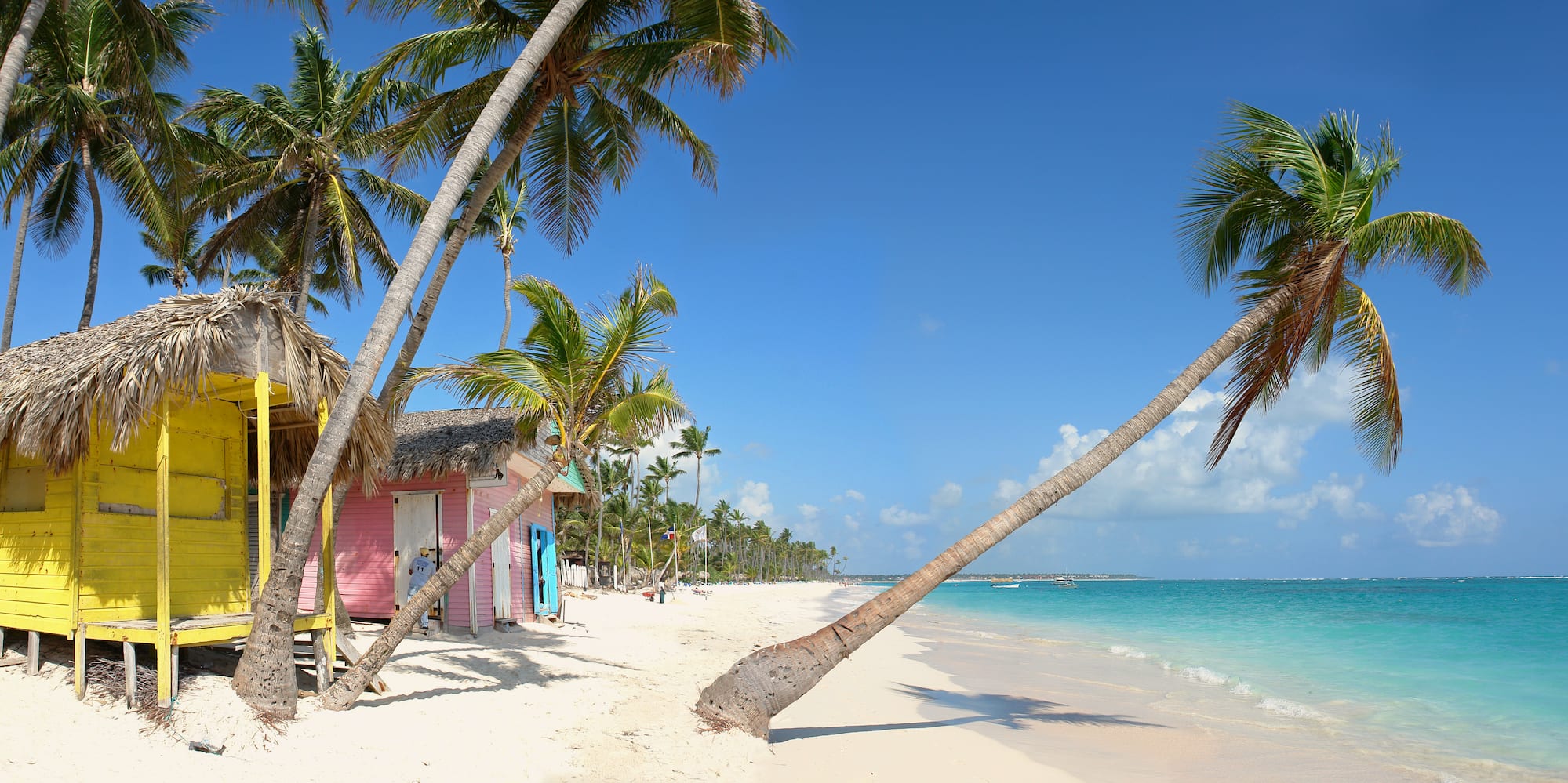Playa tropical con cabañas amarillas y rosas, palmeras y mar turquesa.