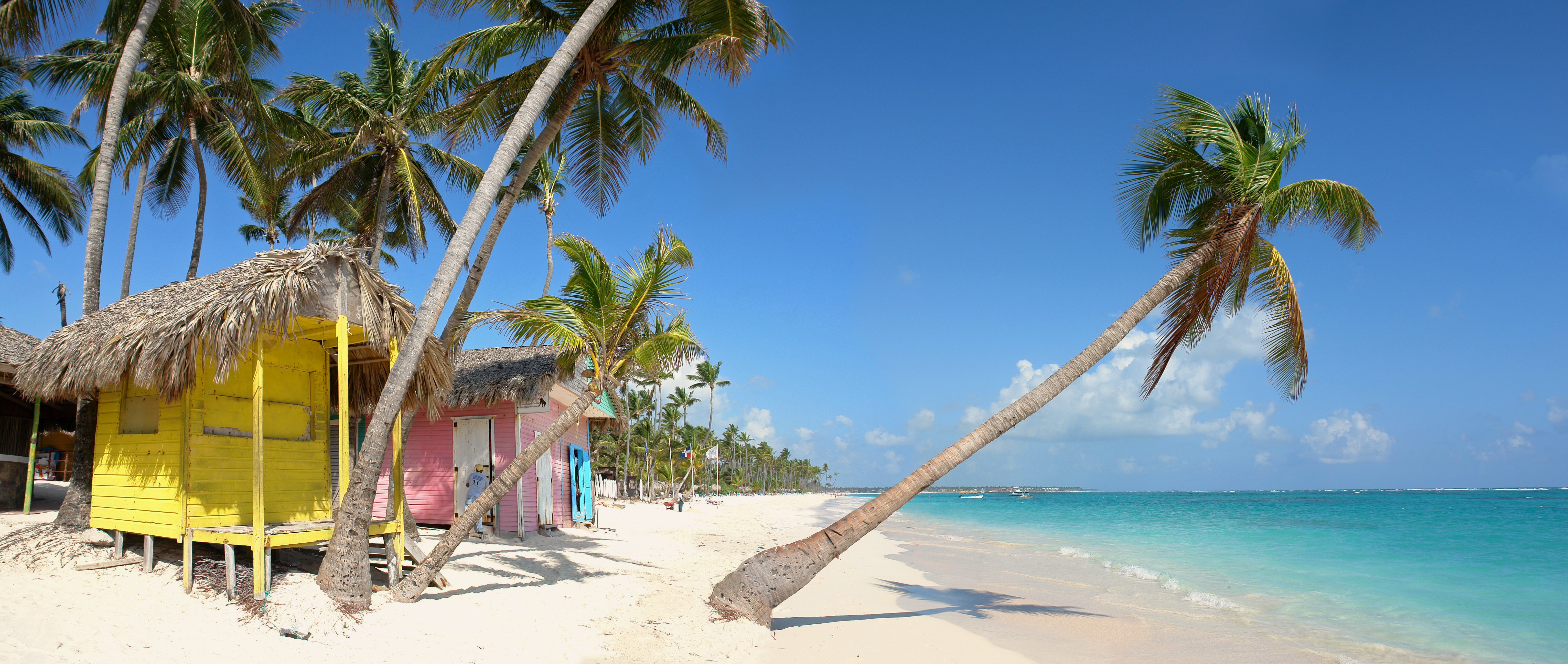 Playa tropical con cabañas amarillas y rosas, palmeras y mar turquesa.