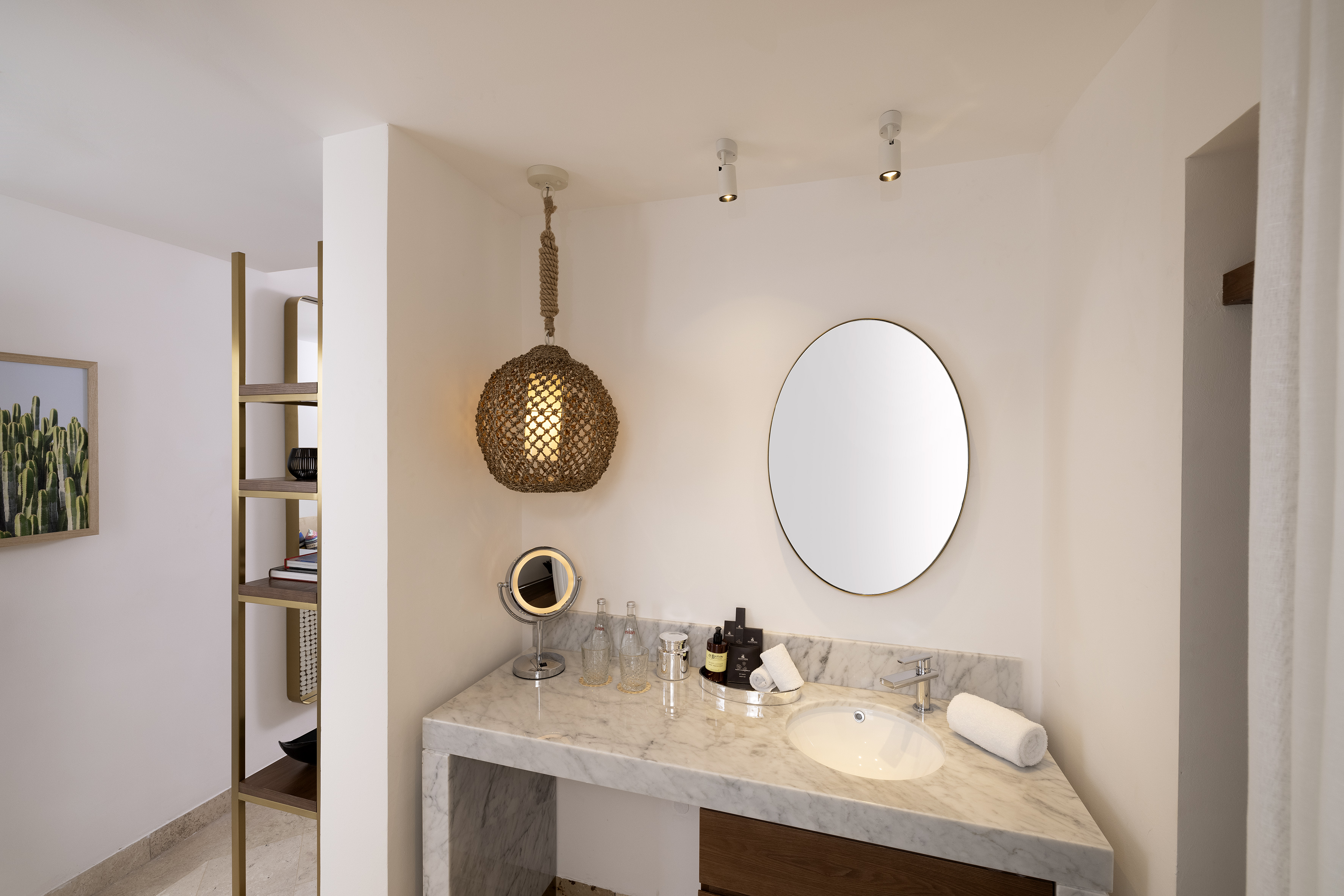 Hotel bathroom with marble vanity, round mirror, woven pendant light, and cactus art.