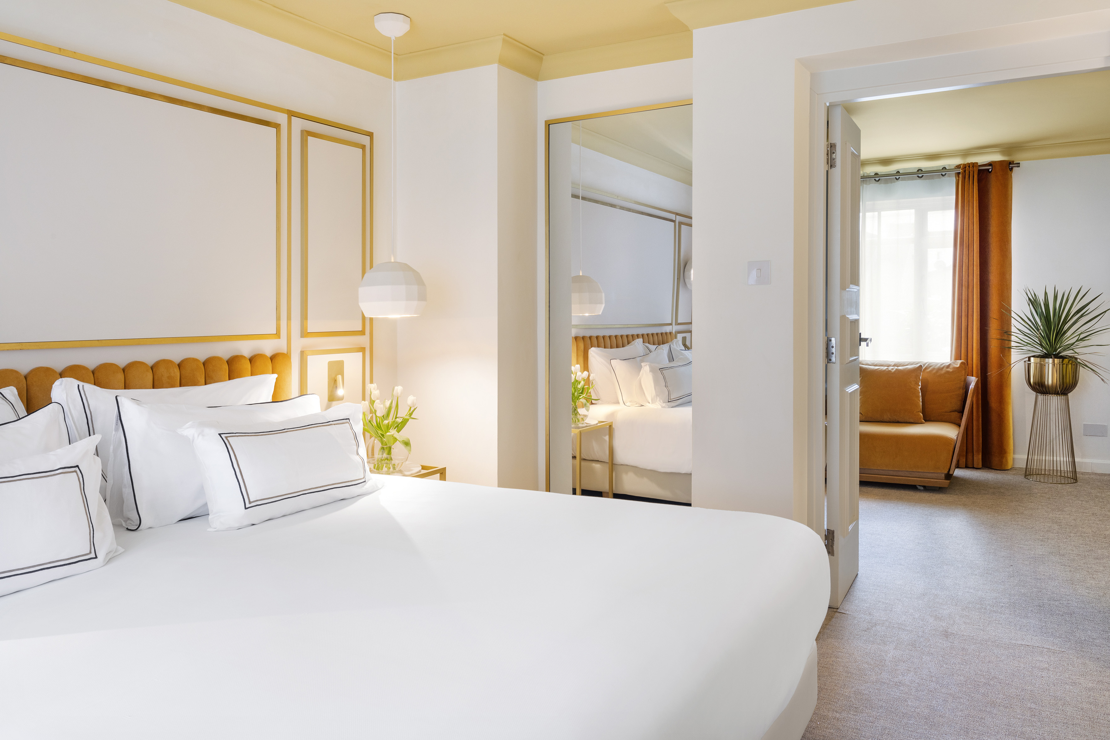Luxury hotel room with white bed, gold details, and view into living area.