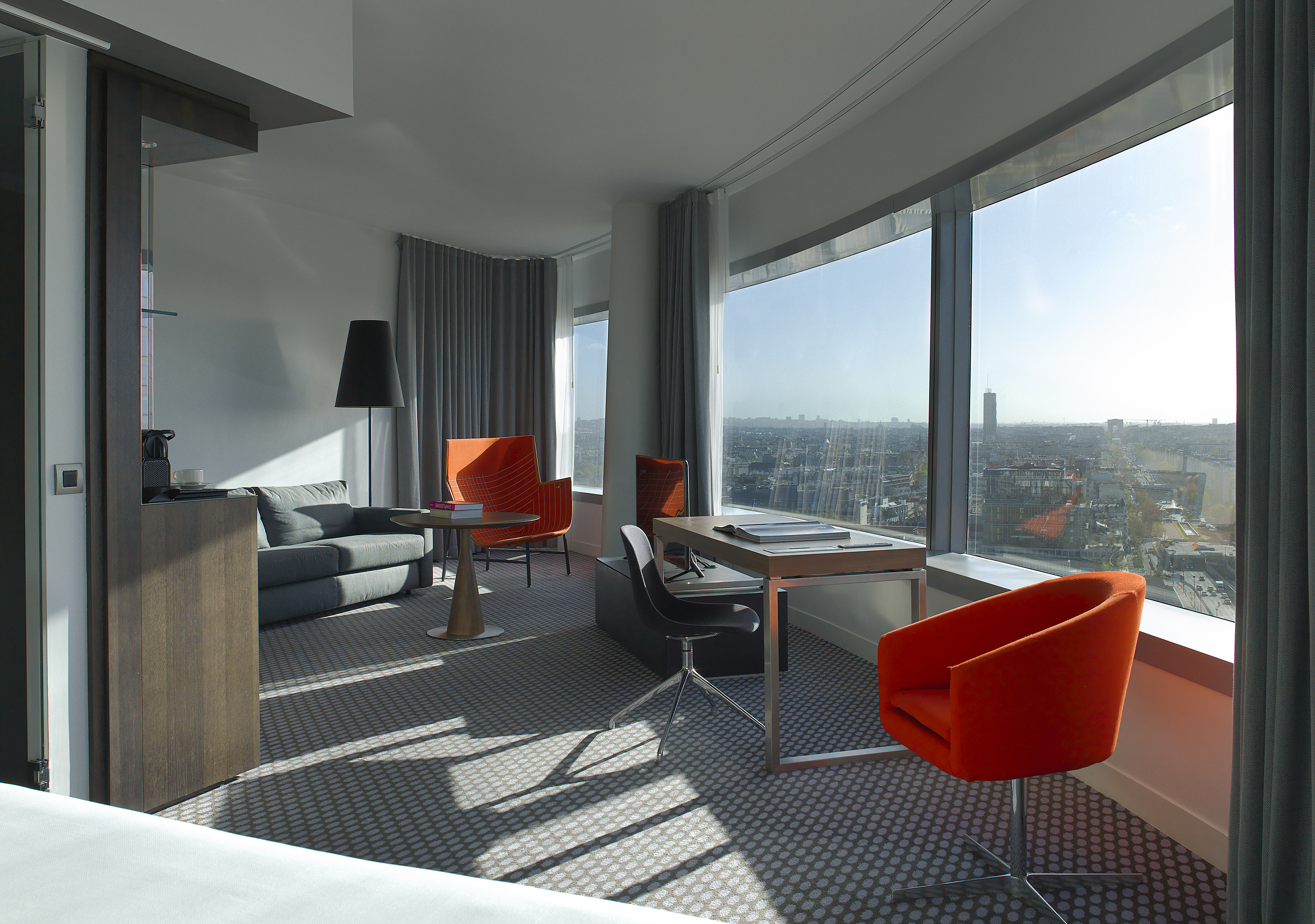 Modern hotel suite, city view, desk, sofa, and orange accent chairs.