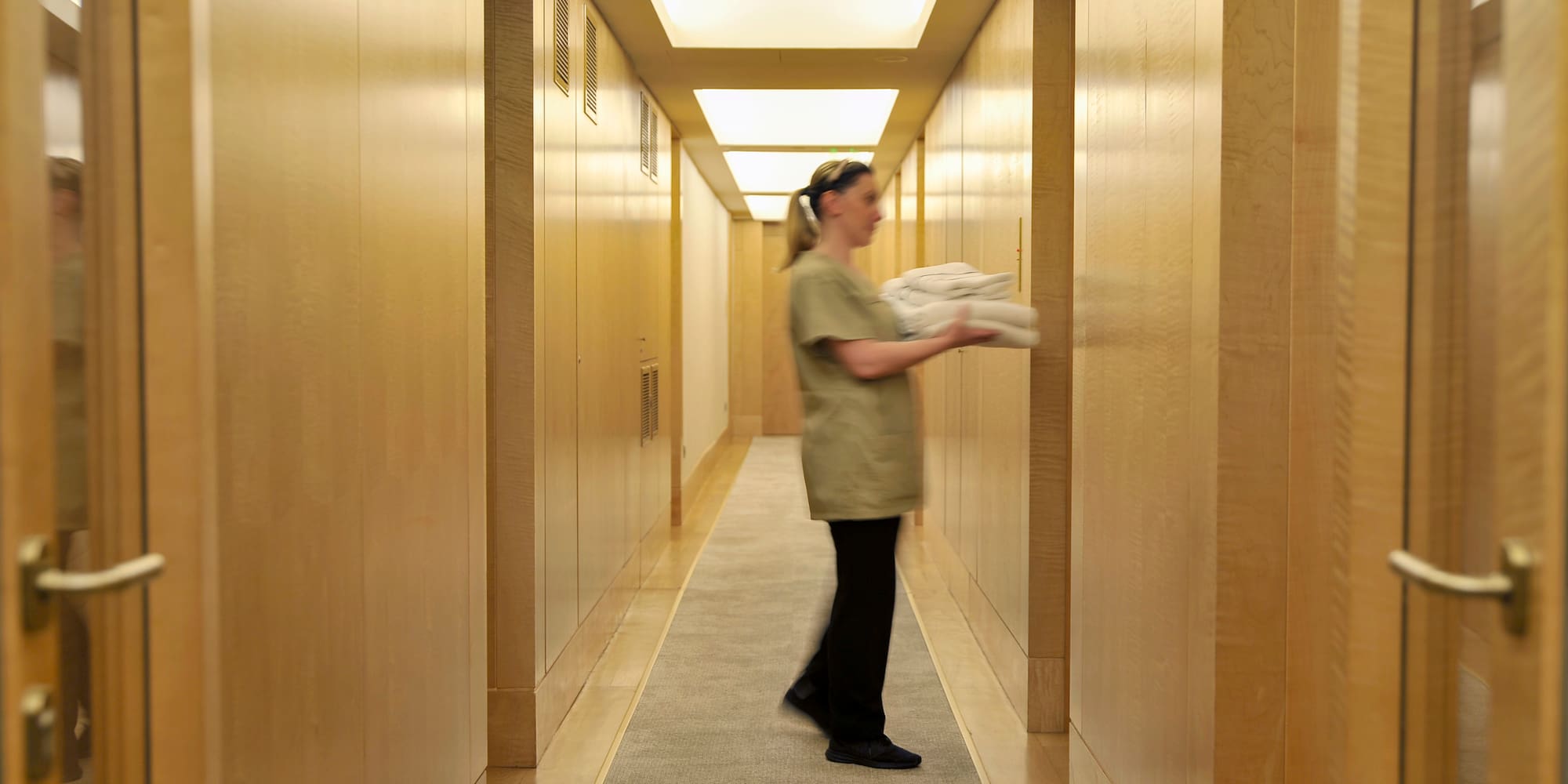 Hotel housekeeper carries fresh towels in a wood-paneled hallway.