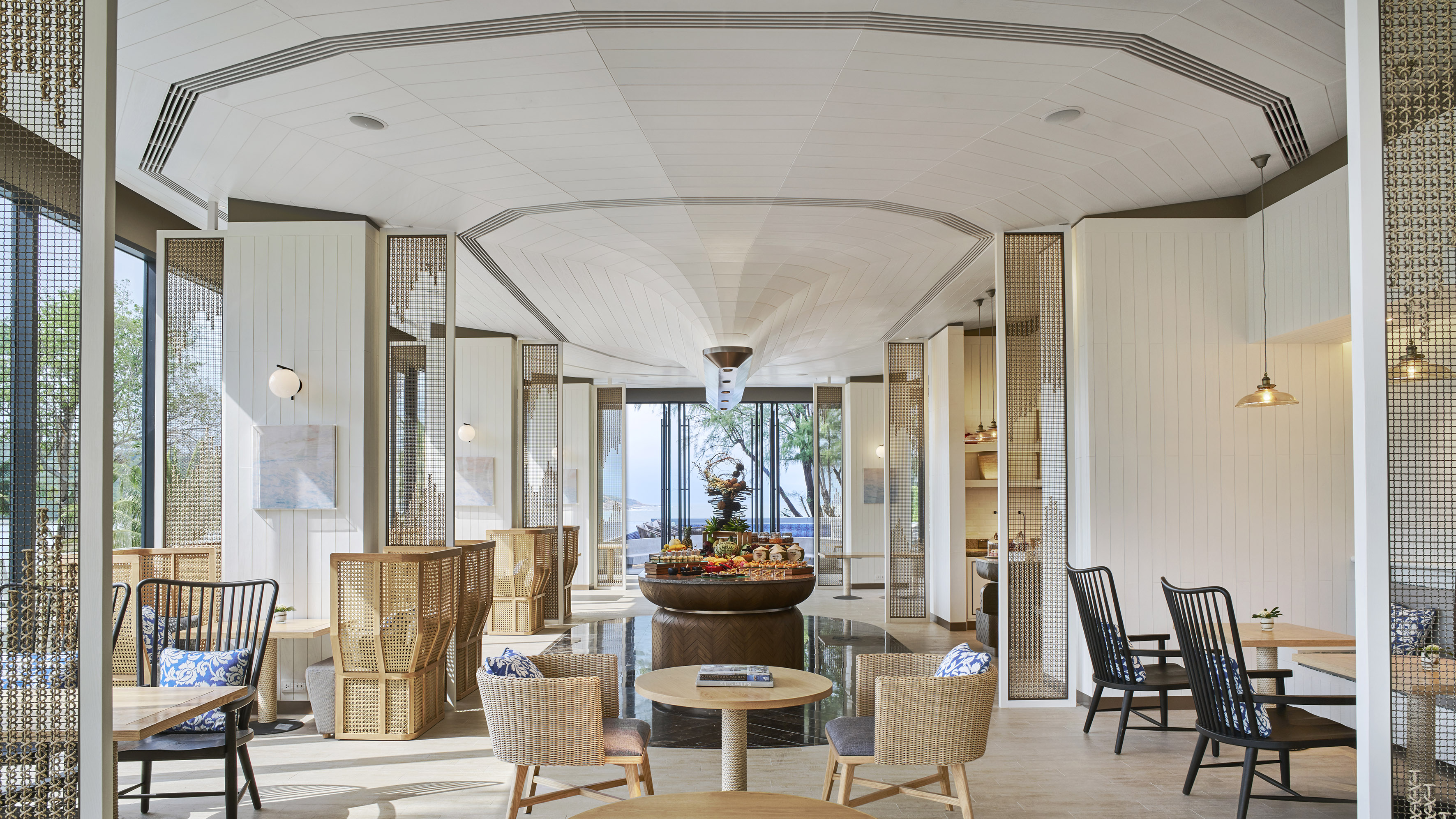 Bright hotel restaurant interior with ocean view, central buffet, and wicker seating.