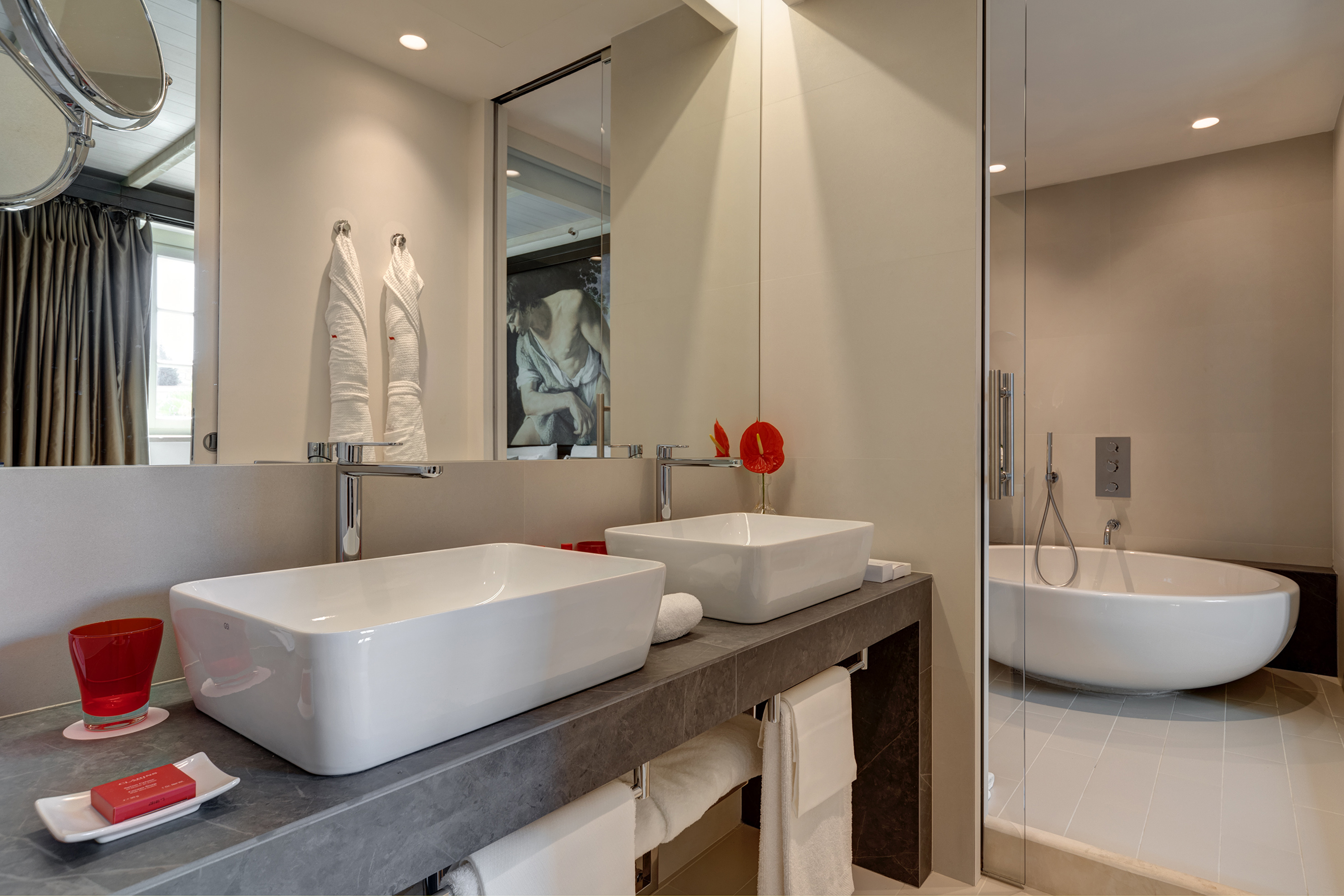Modern hotel bathroom with double white vessel sinks, dark vanity, and freestanding tub.