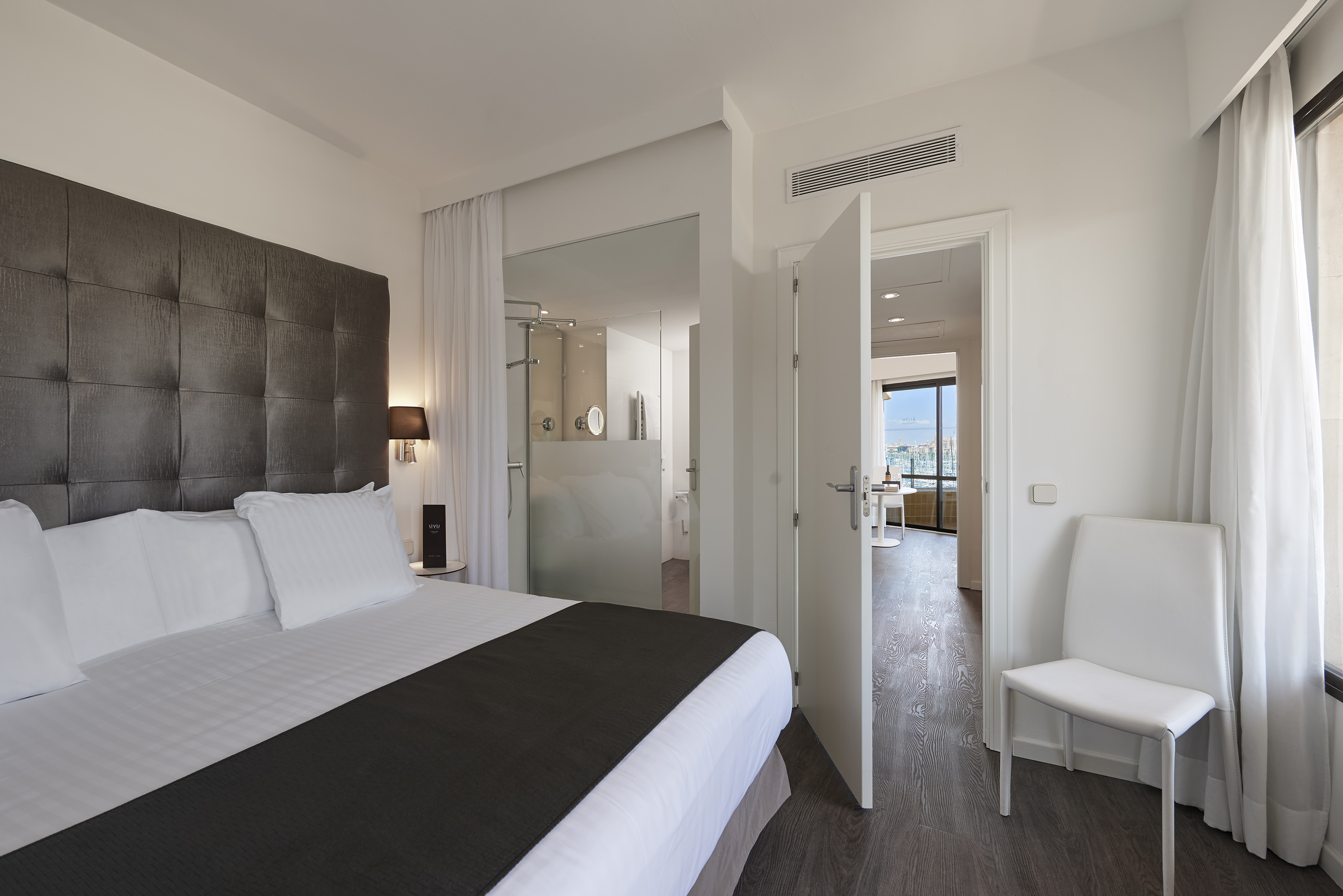 Modern hotel suite bedroom with king bed, glass bathroom, and marina view.