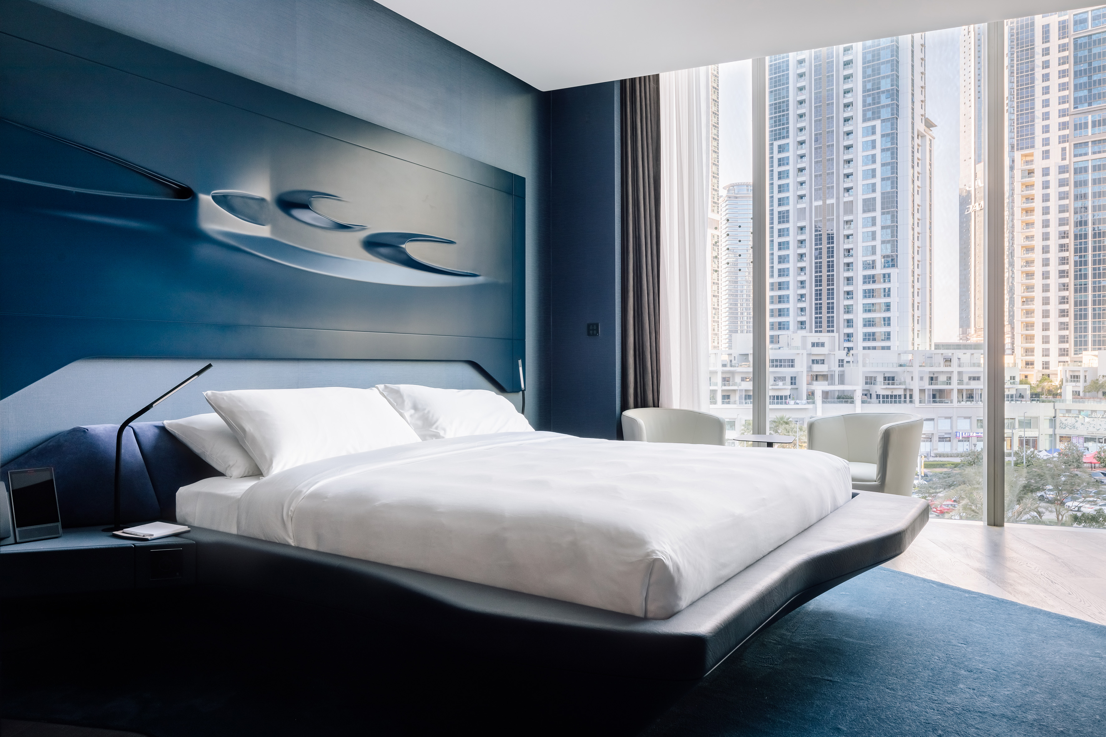 Modern luxury hotel bedroom with blue decor, city skyline view, and large bed.
