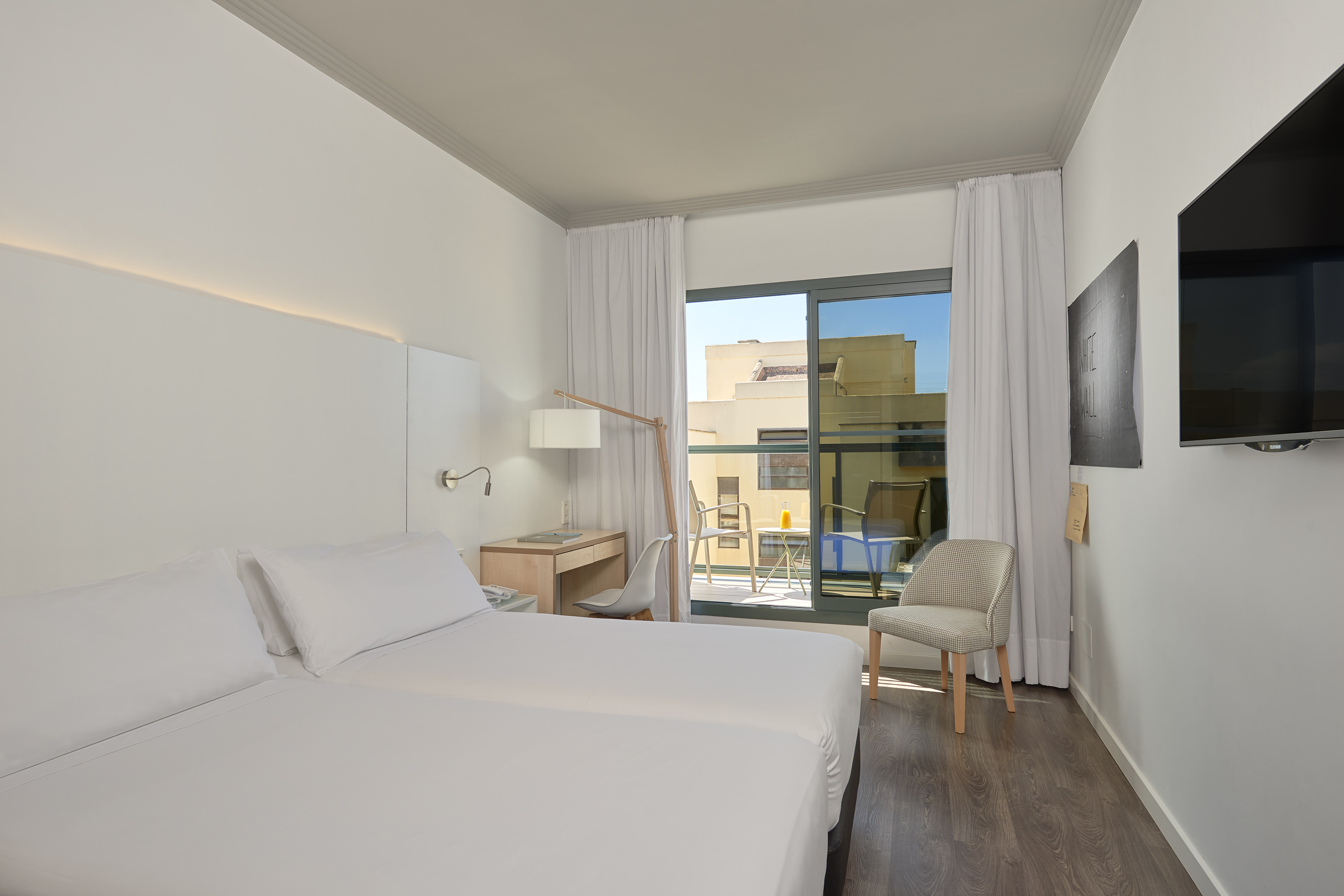 Bright modern hotel room with two beds, a desk, balcony, and city view.