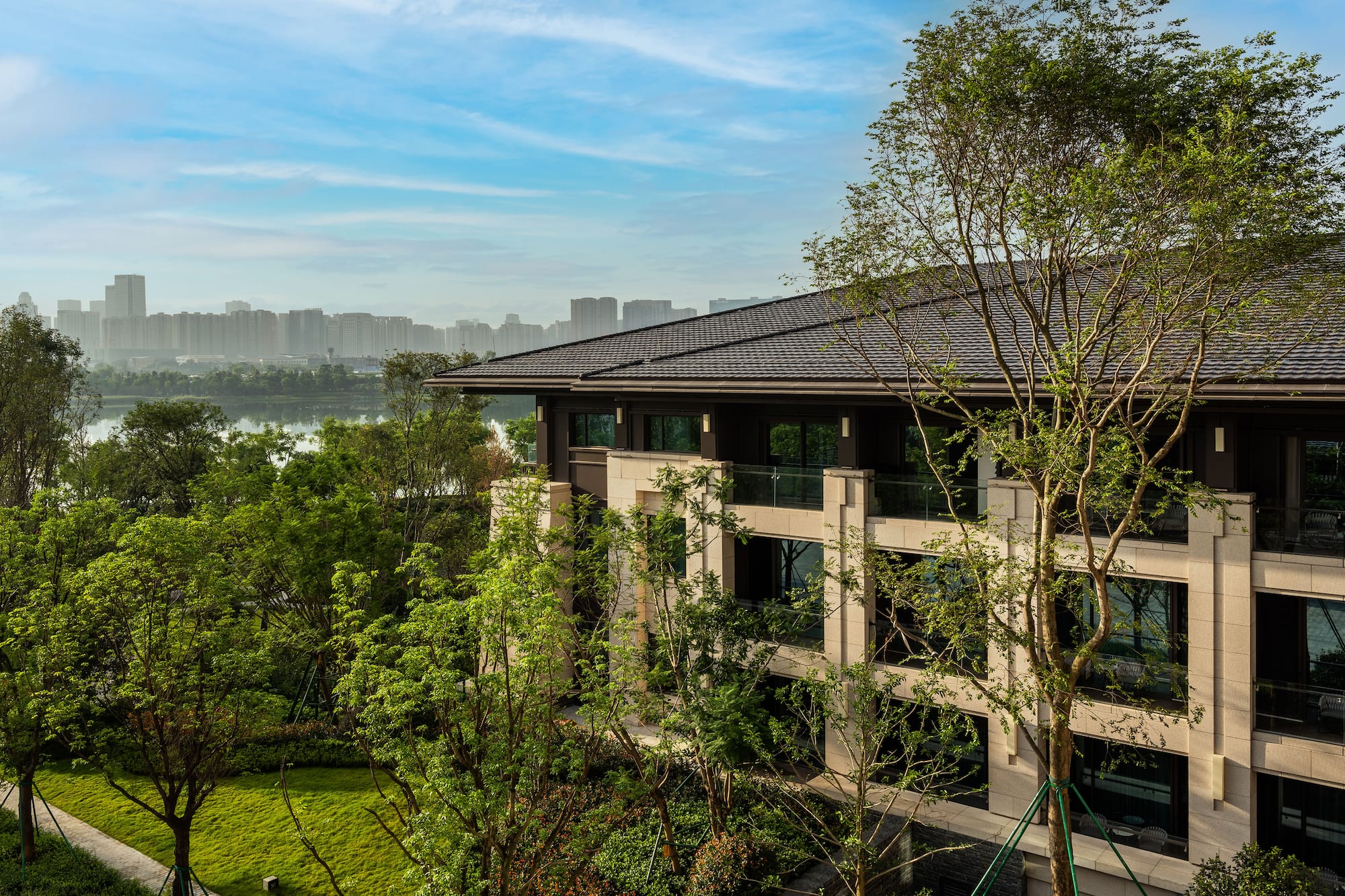 Waterfront modern building with green landscaping and distant city skyline.