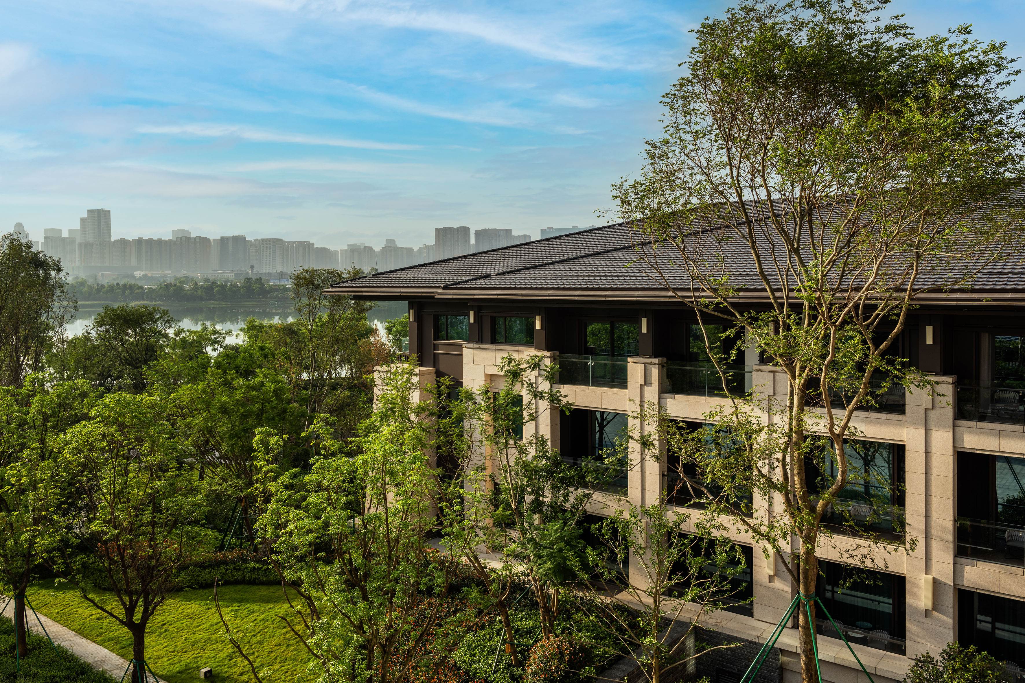 Waterfront modern building with green landscaping and distant city skyline.