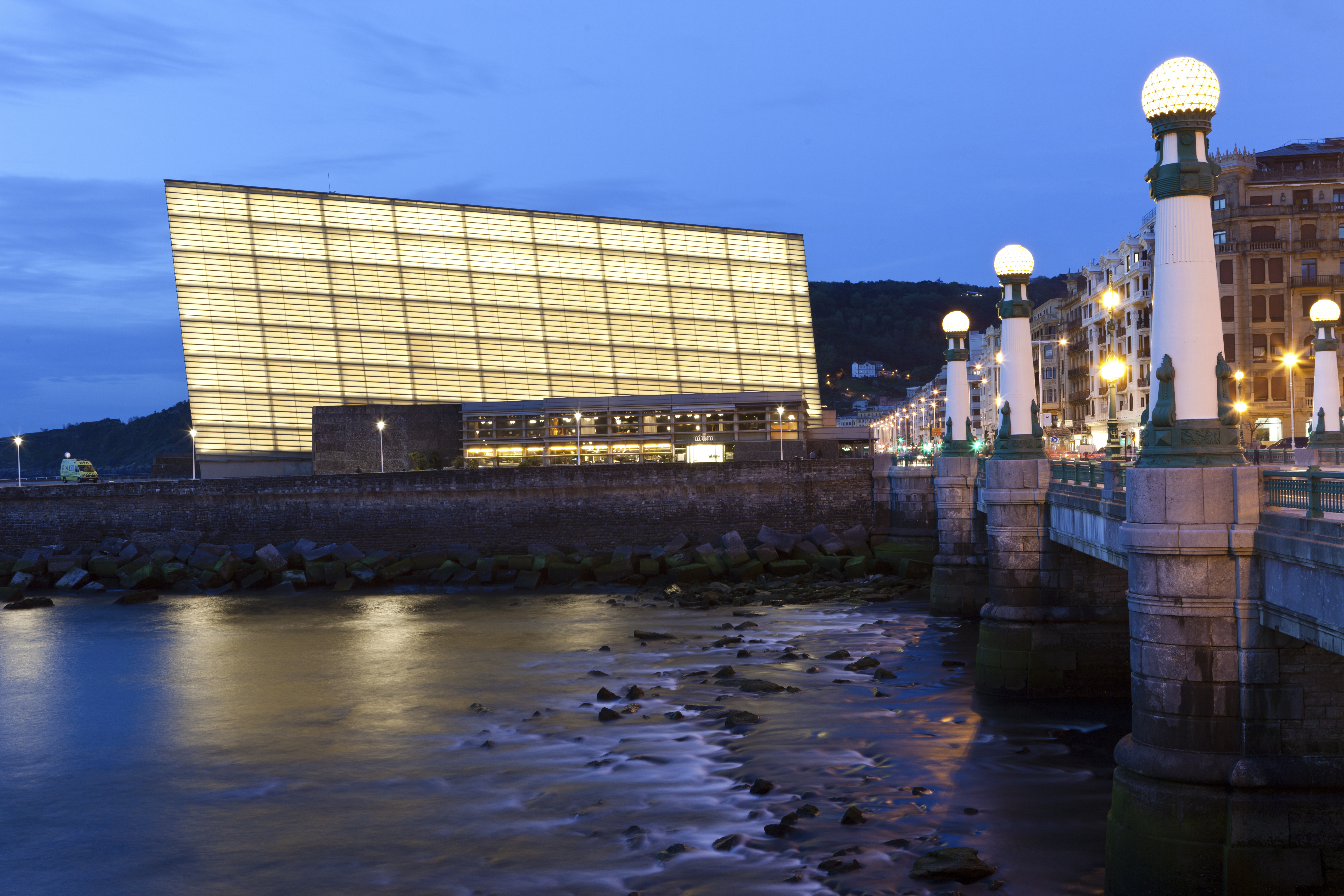 Illuminated Kursaal Center and bridge over river in San Sebastián at dusk.