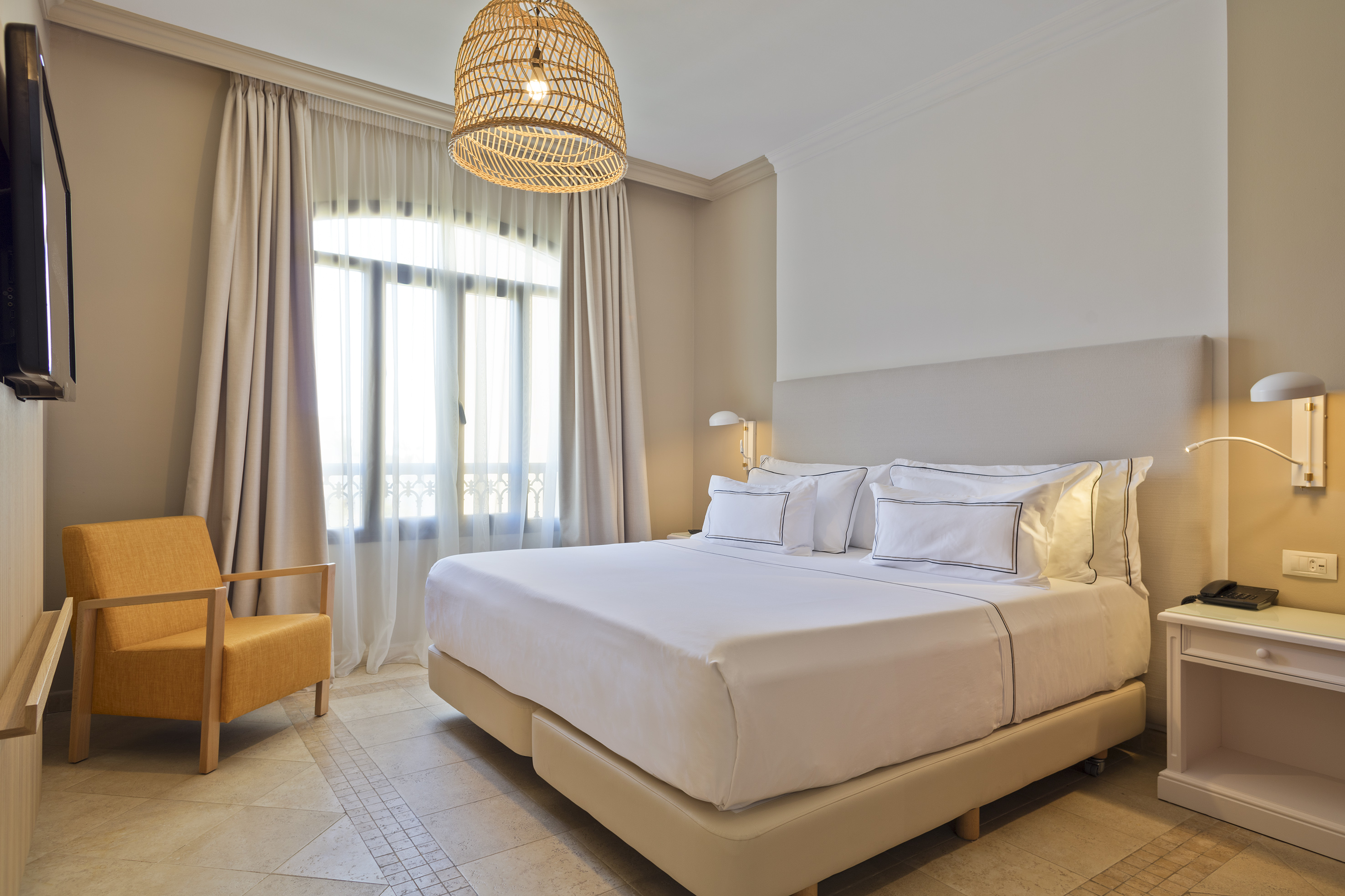 Neutral hotel room: king bed, yellow armchair, window, wicker light.