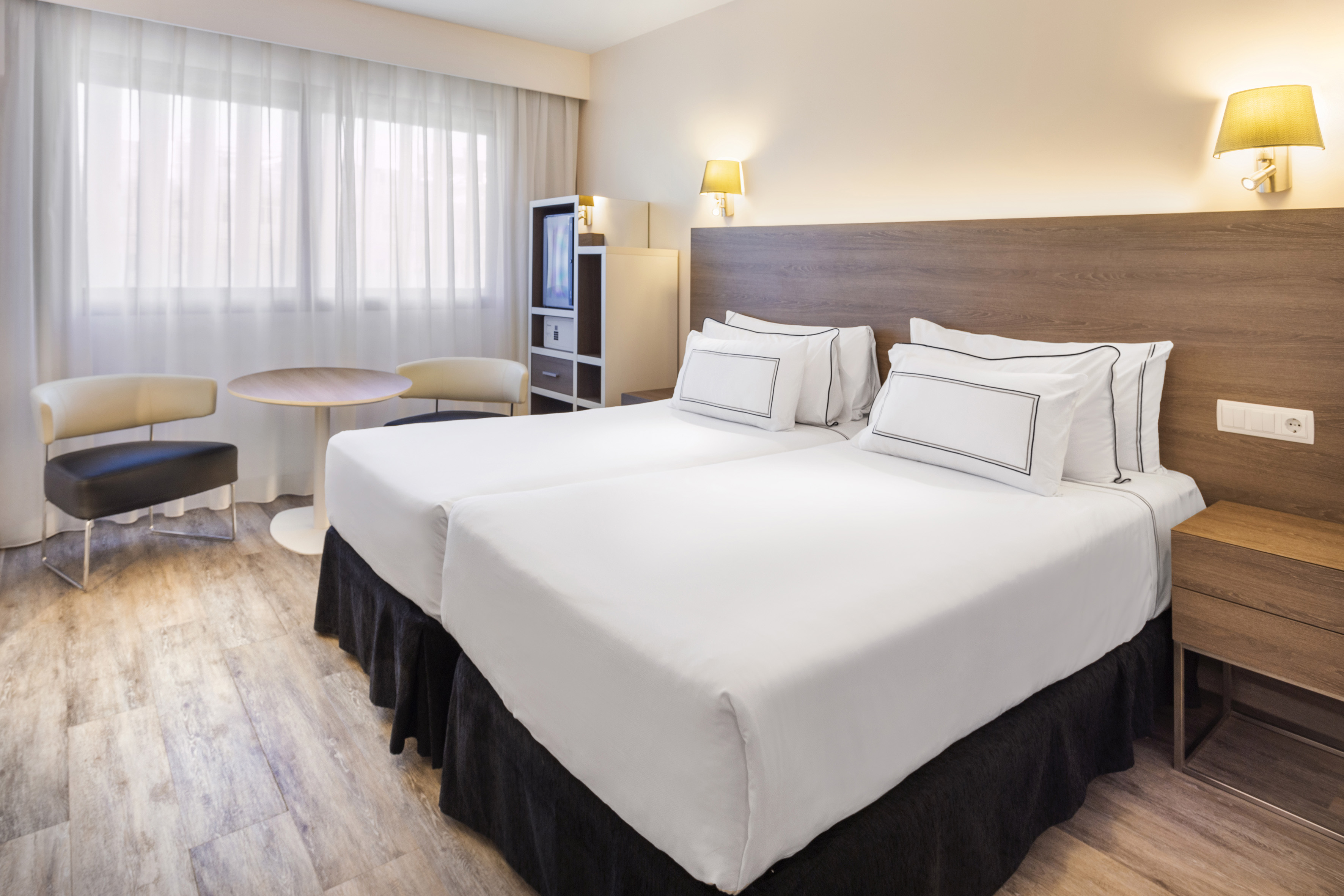 Modern hotel room with two twin beds, wooden headboard, and seating area.