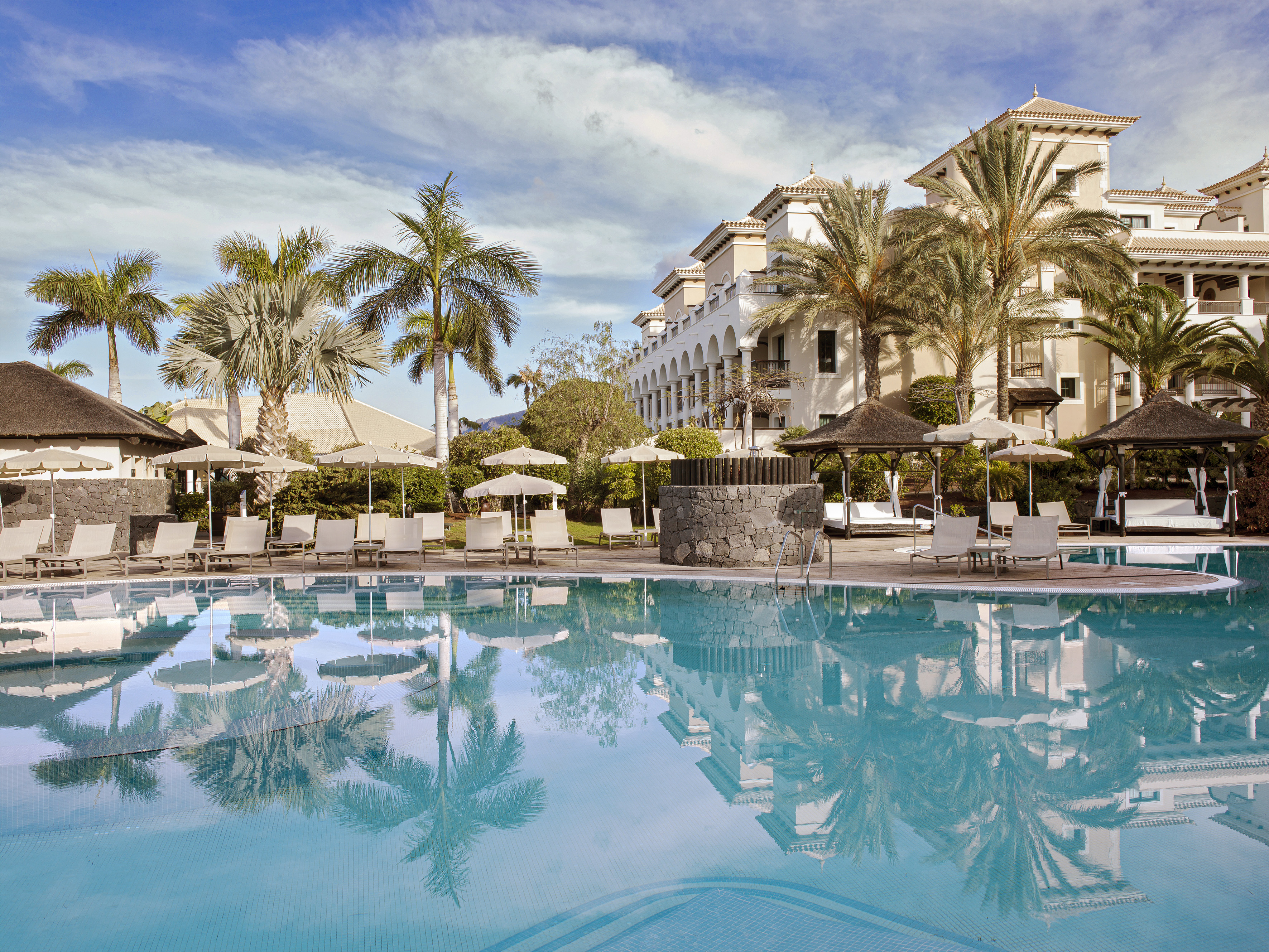Sunny resort pool, palm trees, and Mediterranean-style hotel.