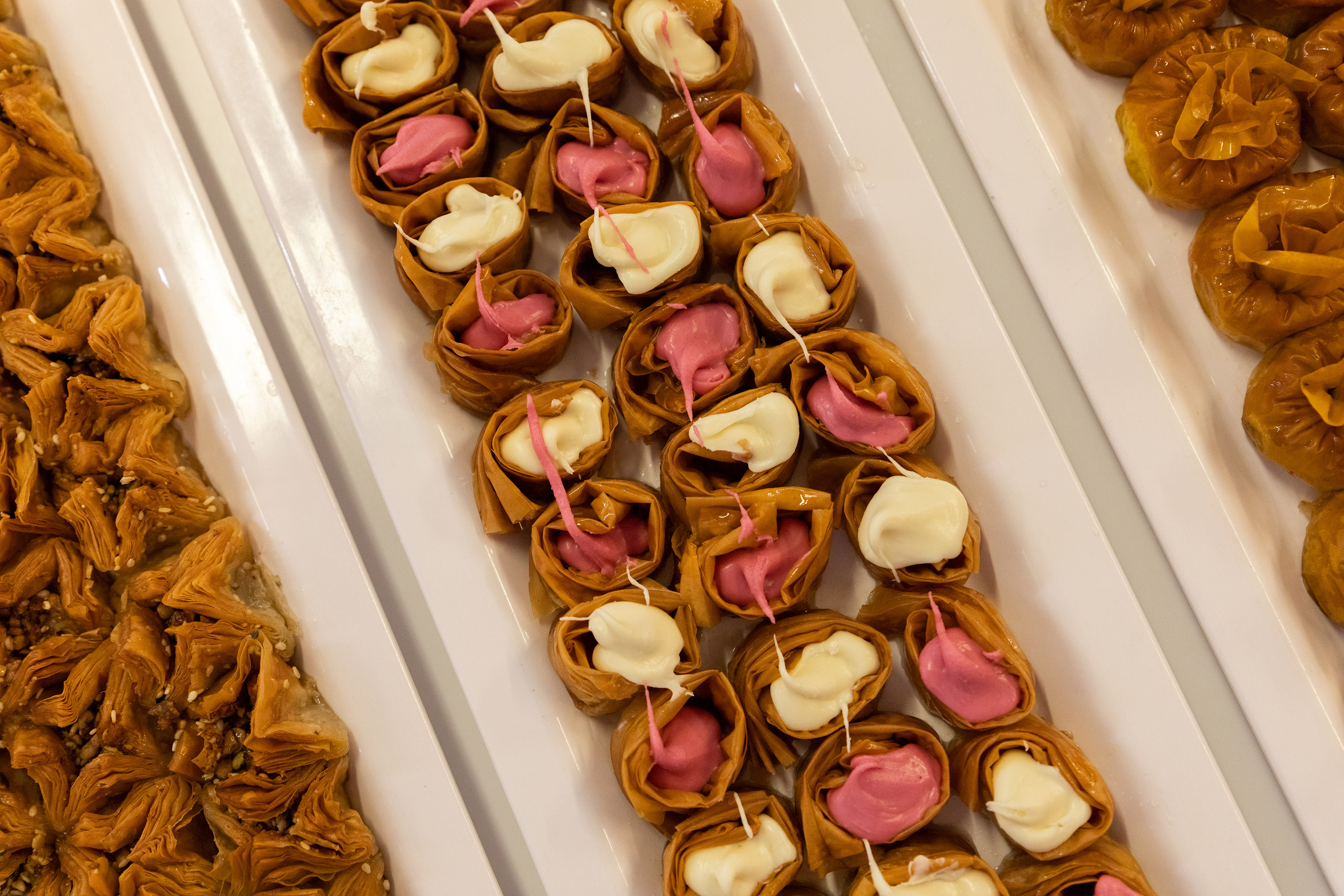 Golden baklava varieties: nut-filled and cream-piped phyllo pastries on white trays.